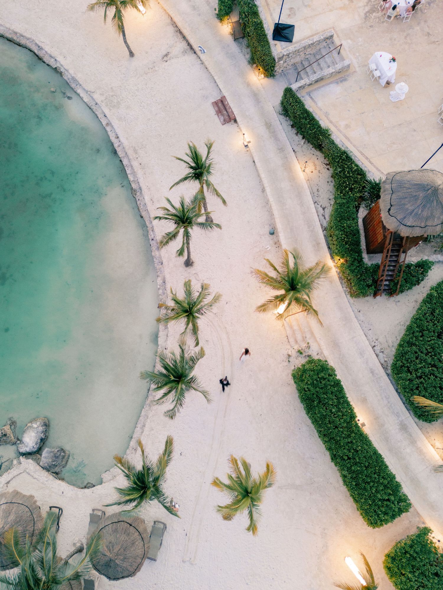Aerial view of a tropical resort with palm trees, sandy paths, turquoise pool, and lush green hedges at dusk.