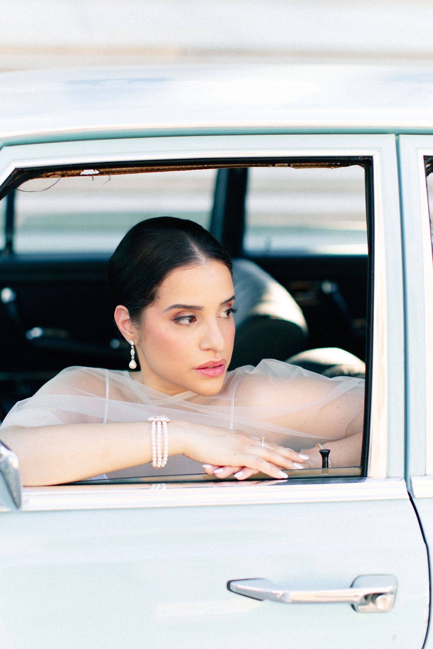 Elegant woman in sheer top leaning out of vintage light blue car window, wearing pearl jewelry.
