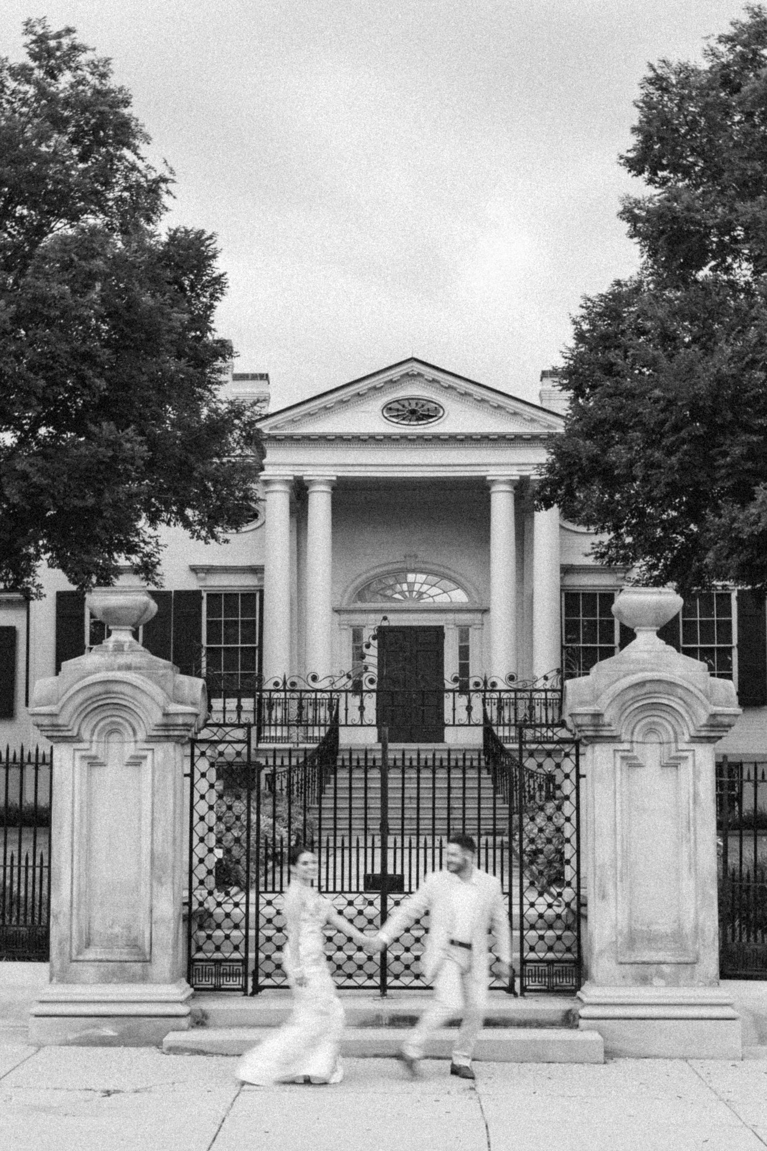 Couple holding hands walking past ornate iron gates of a grand neoclassical building in black and white.