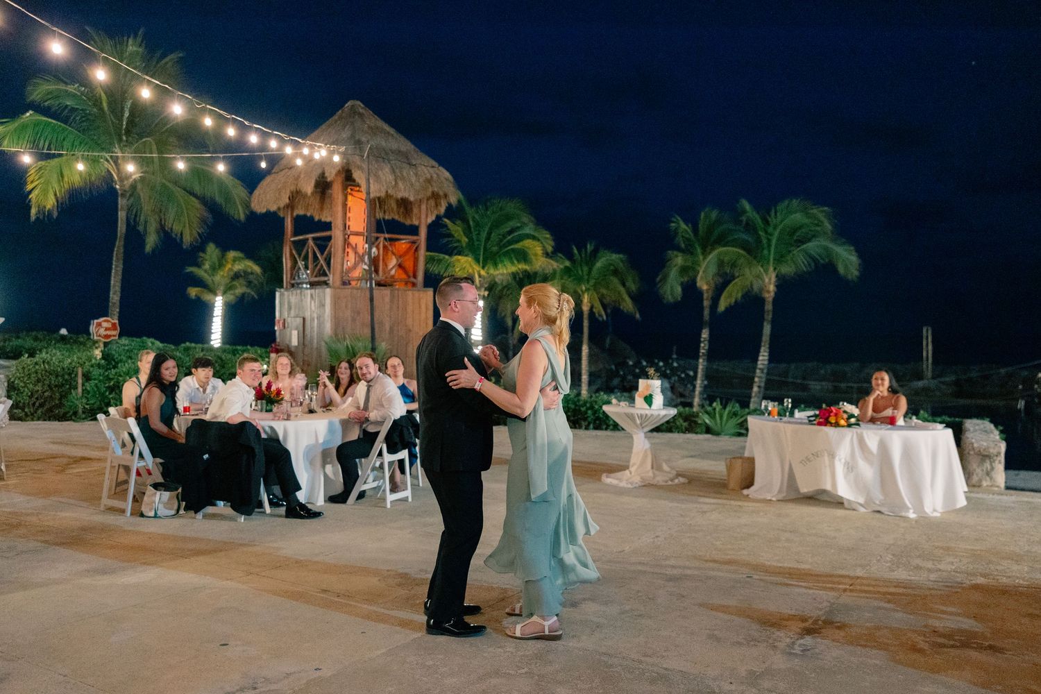 Couple shares first dance at outdoor beach wedding reception at night with string lights and palm trees.