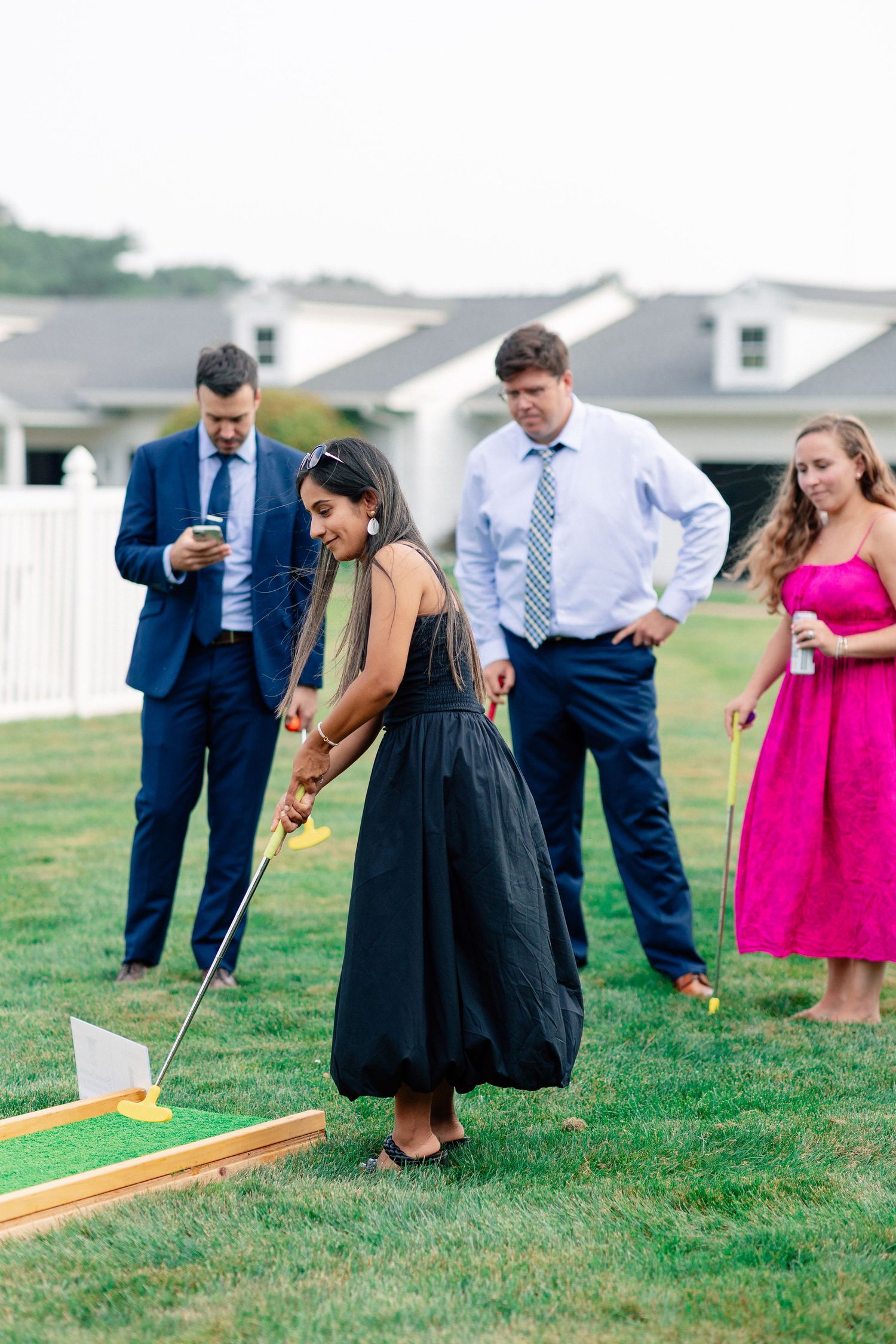 Woman in black dress plays mini golf while guests in formal attire watch at an outdoor lawn event.