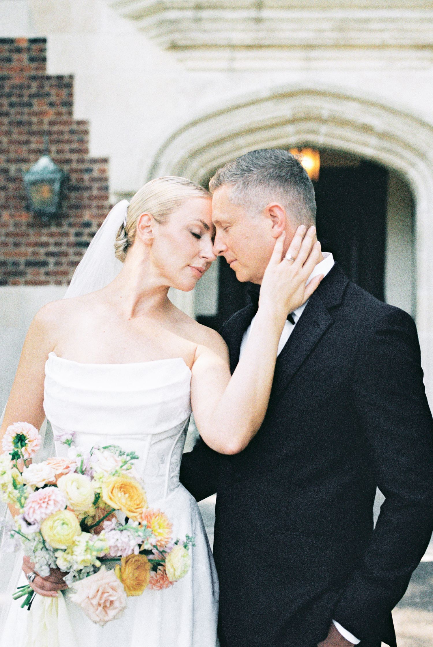 Bride and groom share a tender forehead touch outside a stone archway, bride holding pastel bouquet.