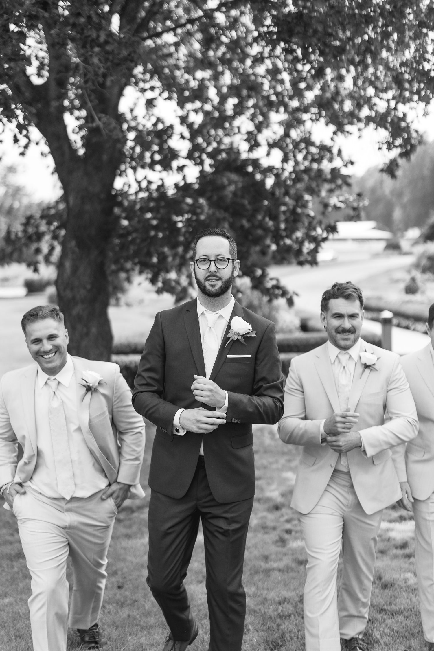 Groom in dark suit leads groomsmen in light suits walking outdoors at wedding ceremony.