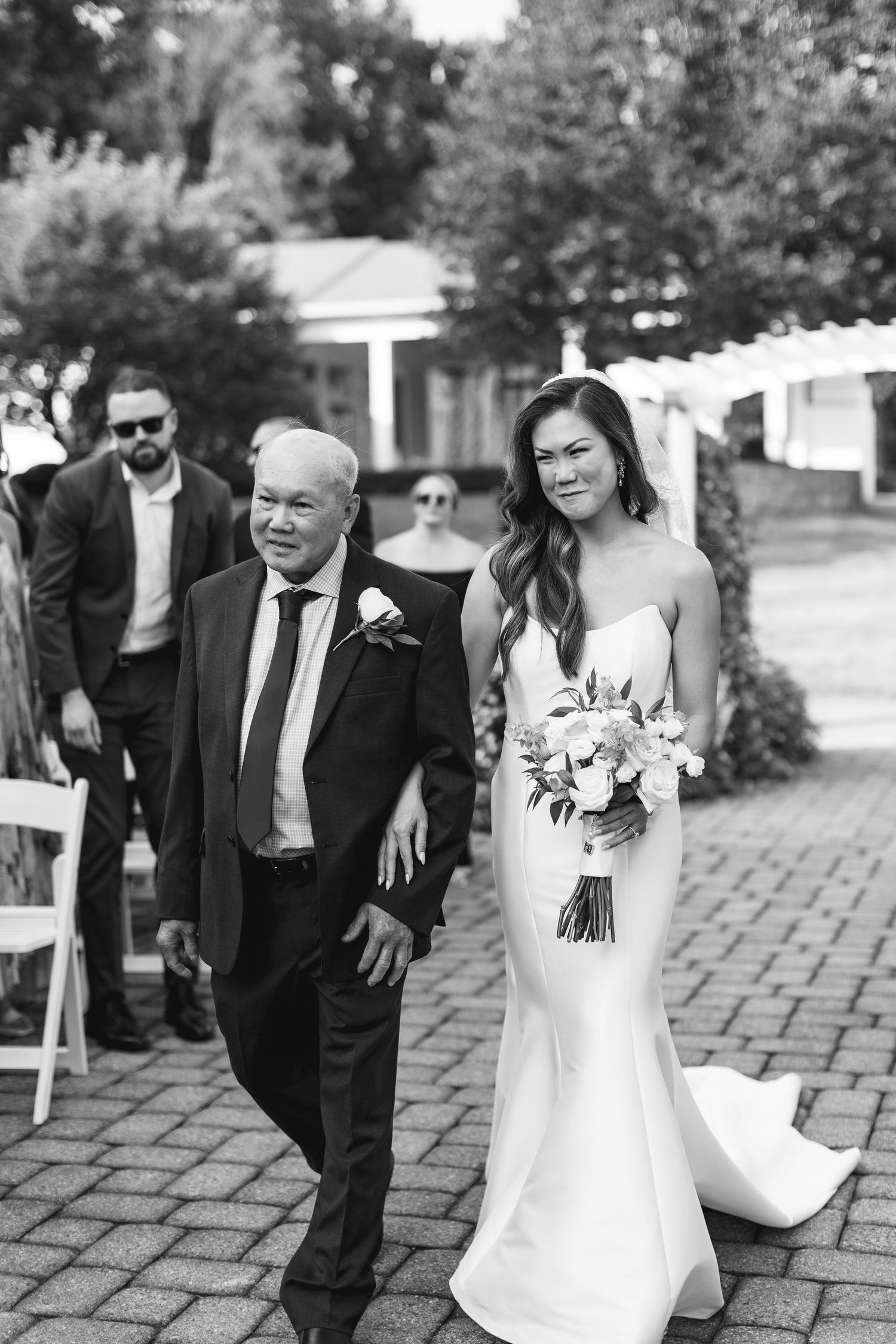 Black and white photo of a bride in a white gown holding a bouquet, walking down the aisle with an older man.