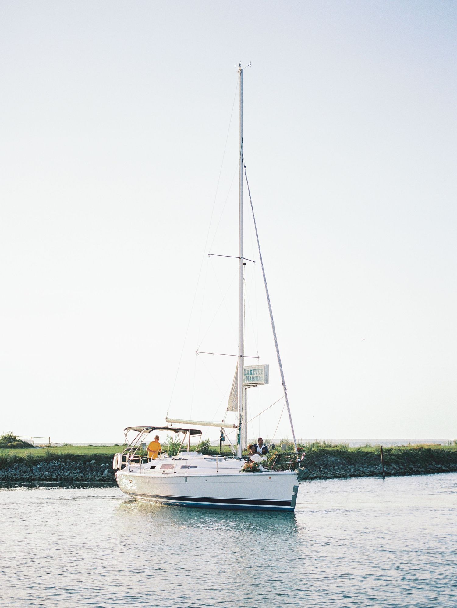 White sailboat with tall mast sailing through calm harbor waters under a clear sky.