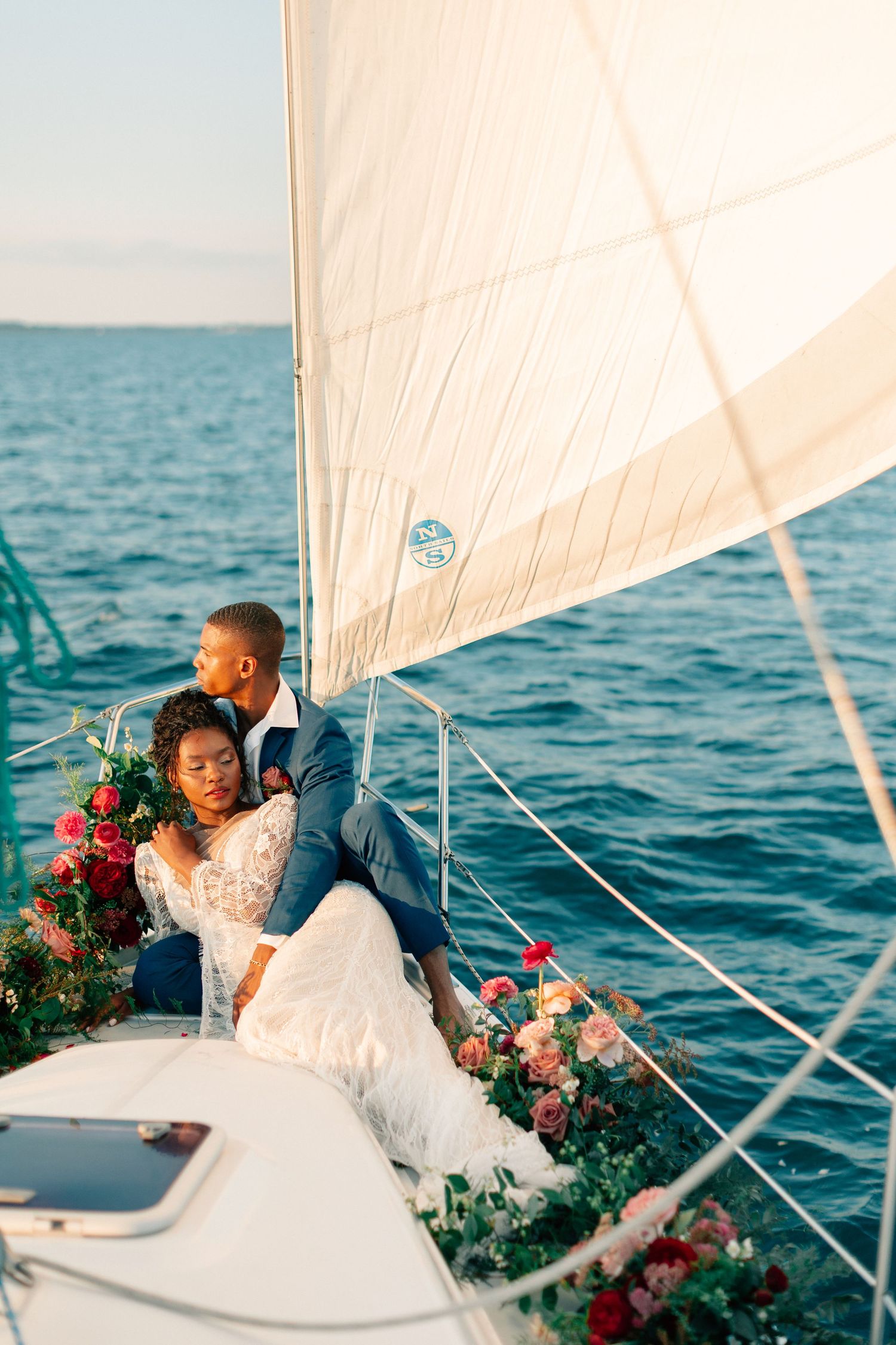 Bride and groom in wedding attire embrace on a sailboat deck surrounded by colorful flowers with ocean views.