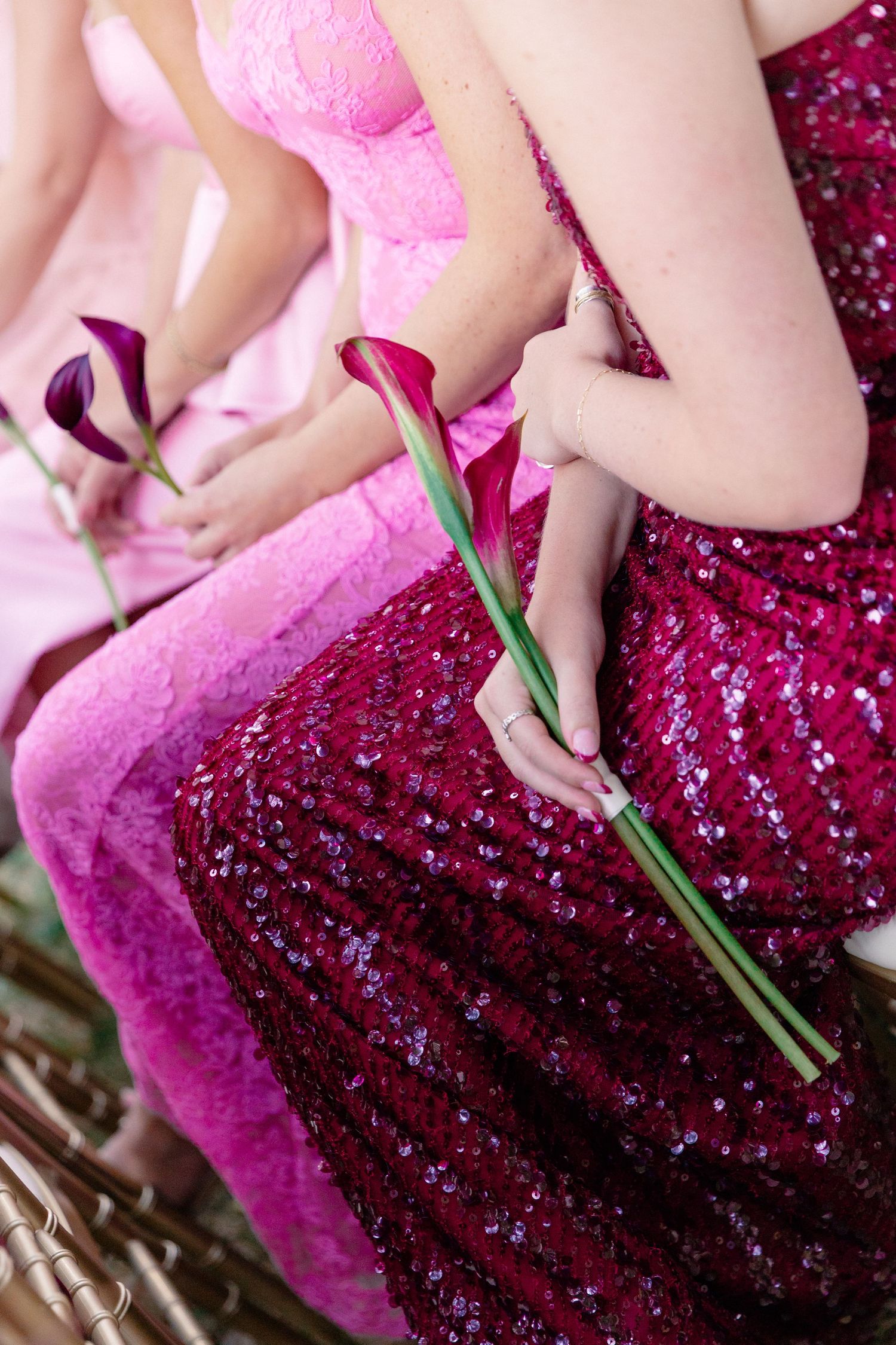 Bridesmaids in pink and magenta sequin gowns hold deep purple calla lily flowers while seated together.
