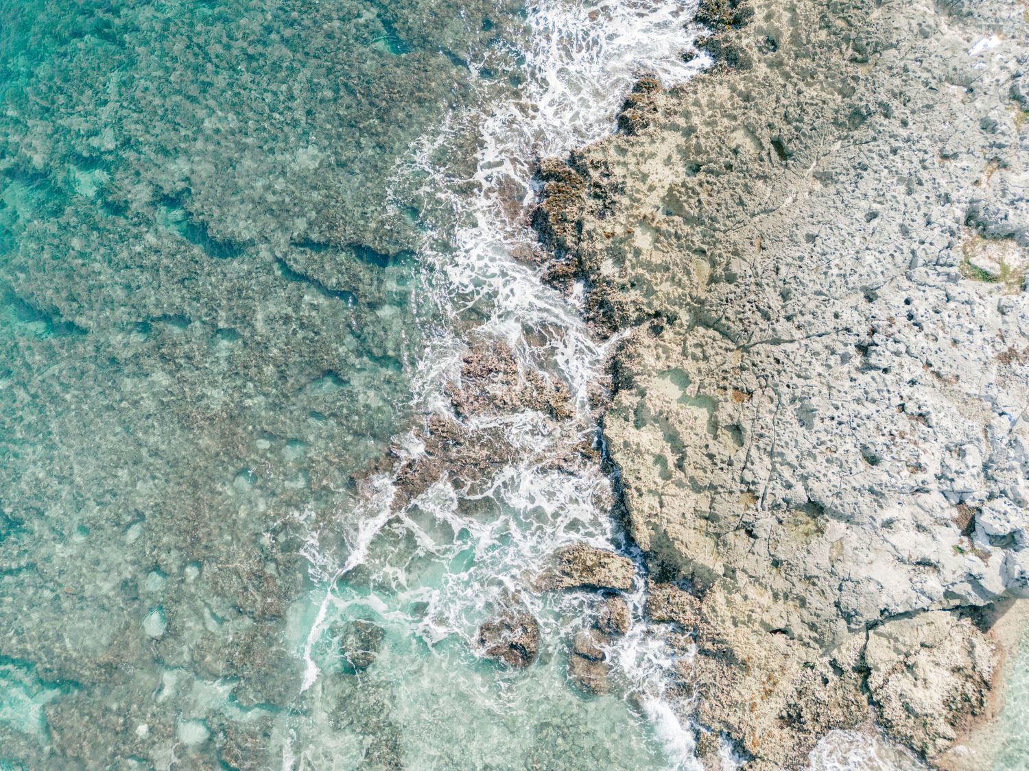 Aerial view of turquoise ocean waves washing over rocky coral reef in clear tropical waters.