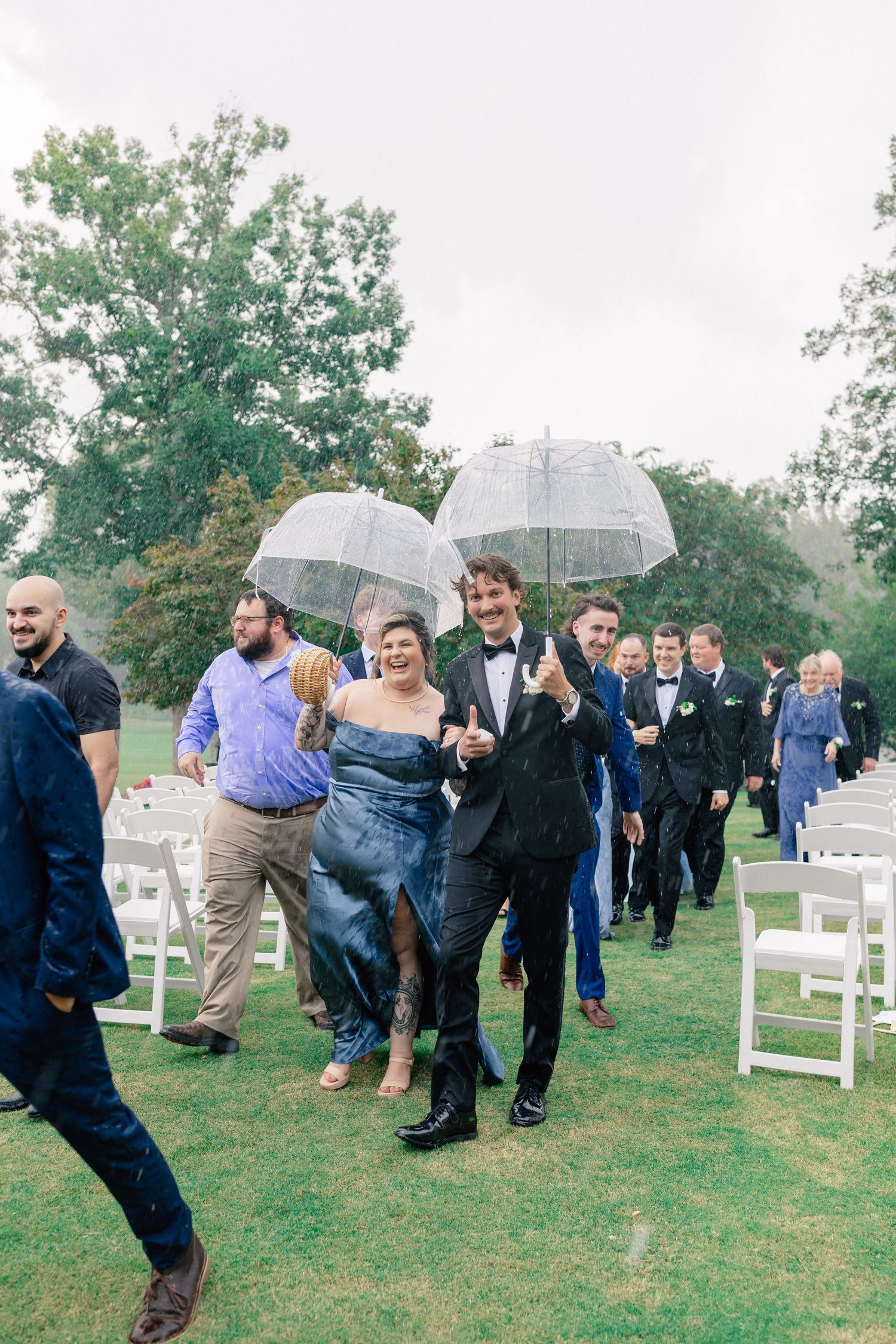 Wedding party walks down outdoor aisle holding clear umbrellas in the rain after ceremony.