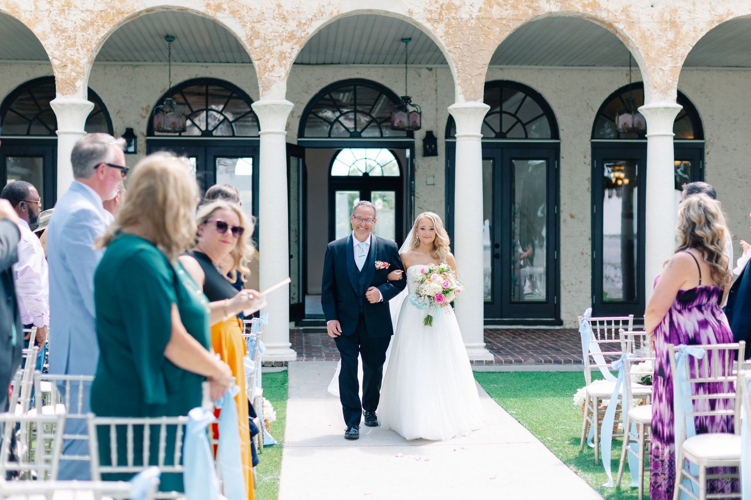 Bride walks down outdoor aisle with escort at elegant venue wedding ceremony, guests standing alongside white chairs, wedding at Bella Cosa