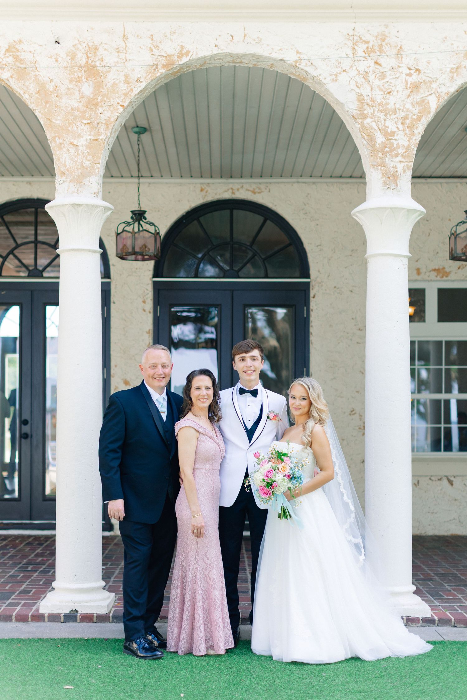 Family poses with bride and groom in white tuxedo before elegant arched building entrance on wedding day, wedding at Bella Cosa