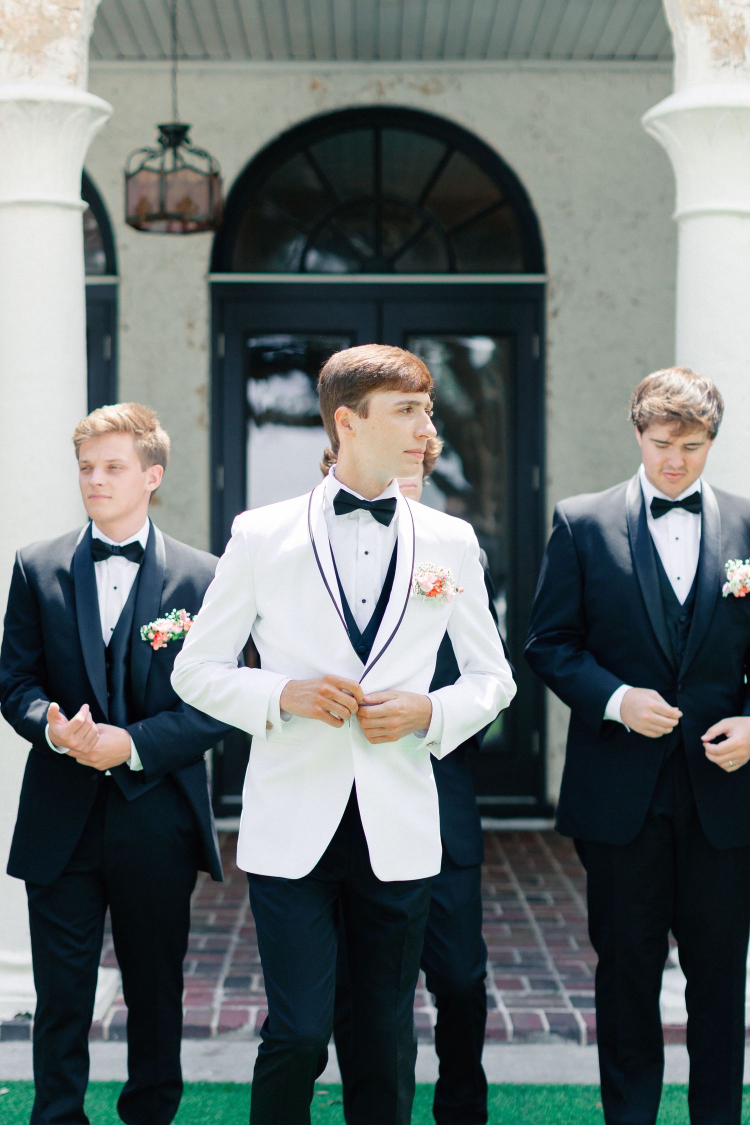 Groom in white tuxedo jacket stands with two groomsmen in black tuxedos outside elegant venue, wedding at Bella Cosa