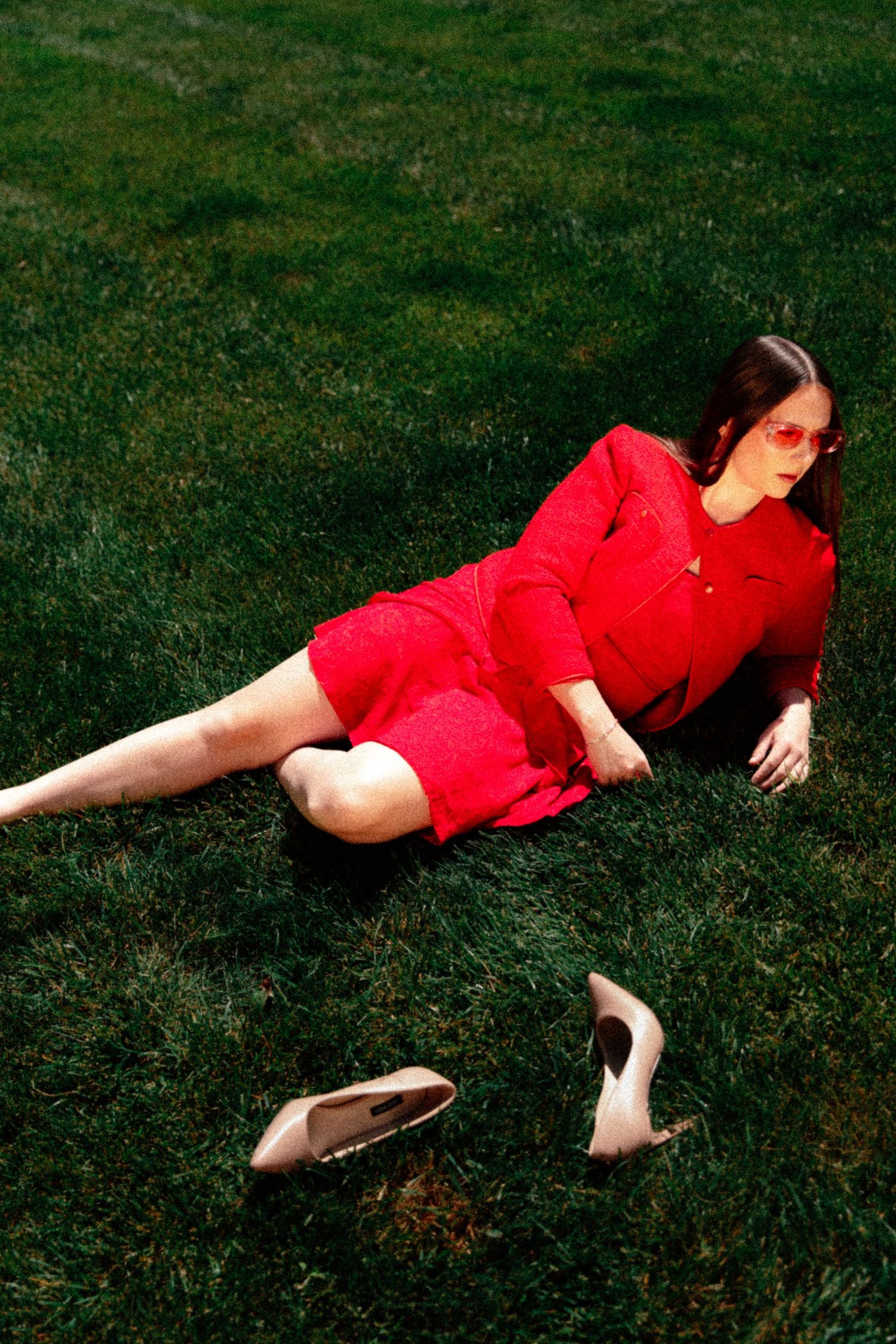 Woman in red dress and sunglasses lying on green grass with nude heels nearby.