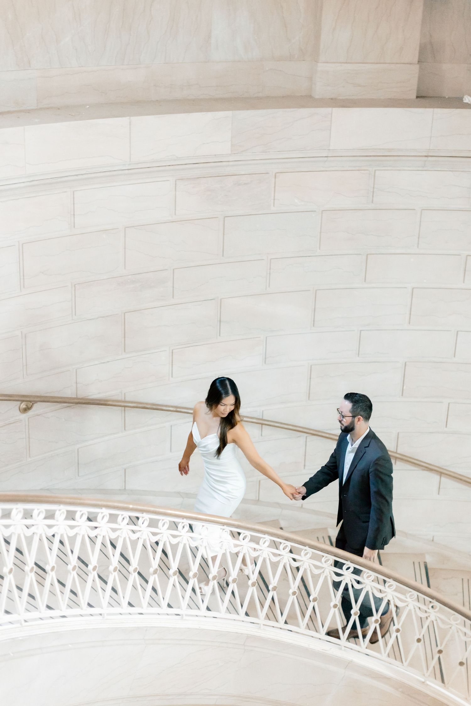 Couple holds hands walking up elegant spiral staircase with ornate white railing in a grand marble building.