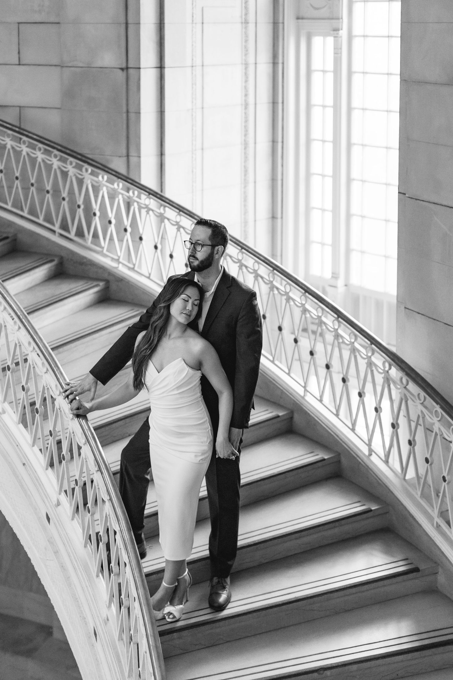 Black and white photo of a couple embracing on an elegant curved staircase, woman in white dress, man in dark suit.