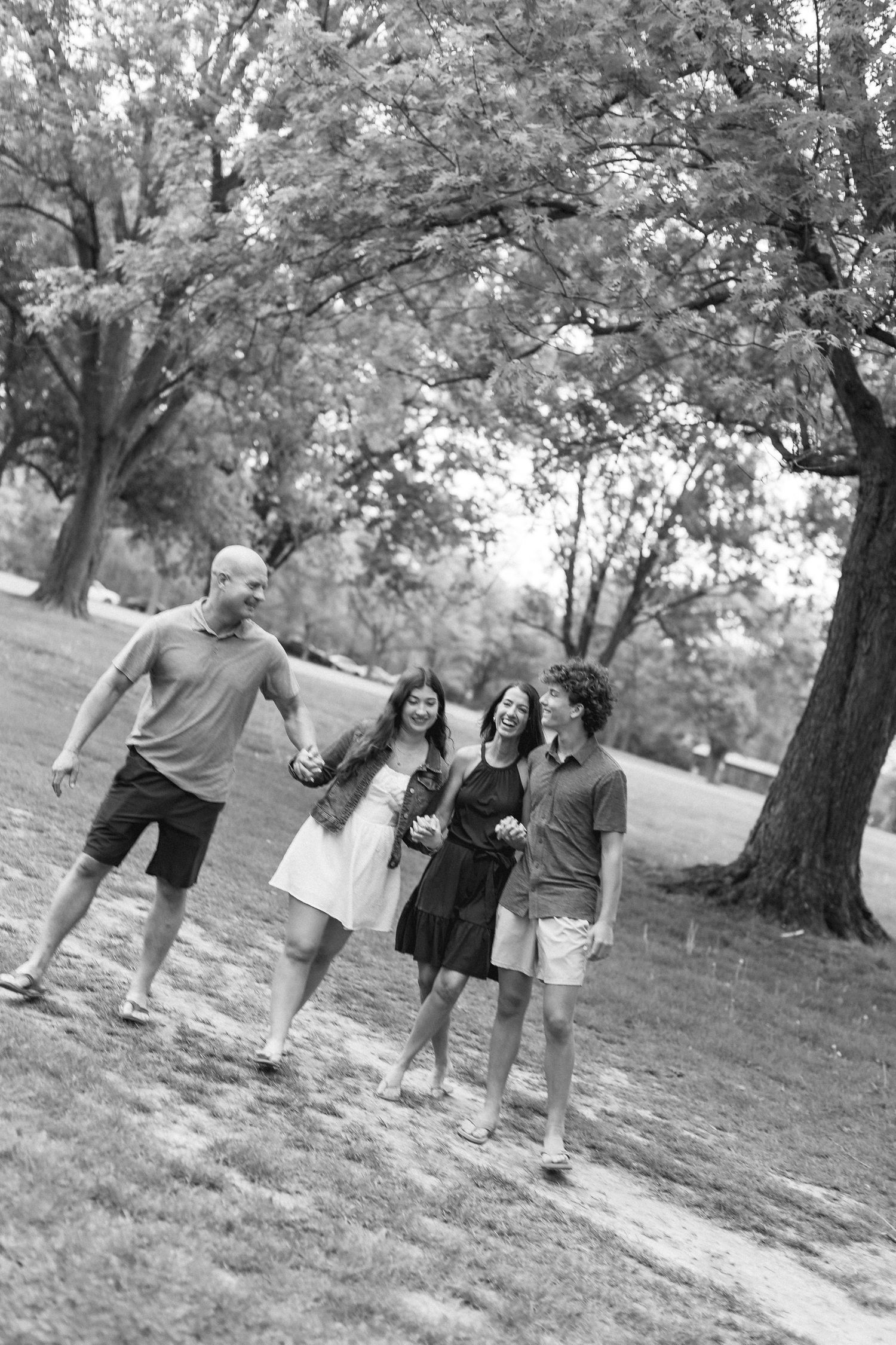 Black and white photo of a happy family of four walking together in a lush park, laughing and holding hands.