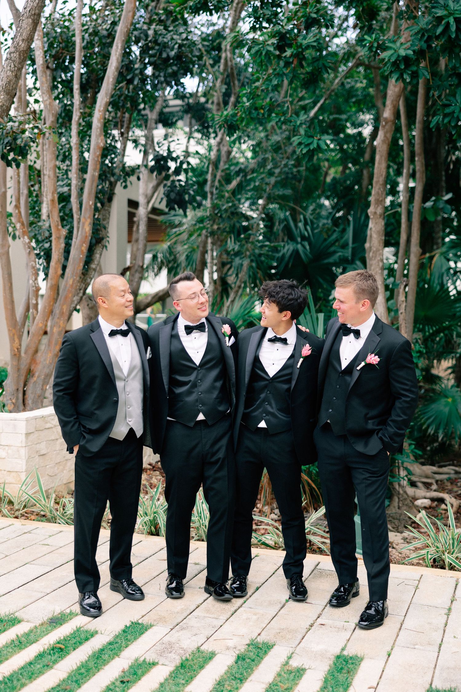 Four groomsmen in black tuxedos with bow ties and boutonnieres laughing together outdoors at a wedding.