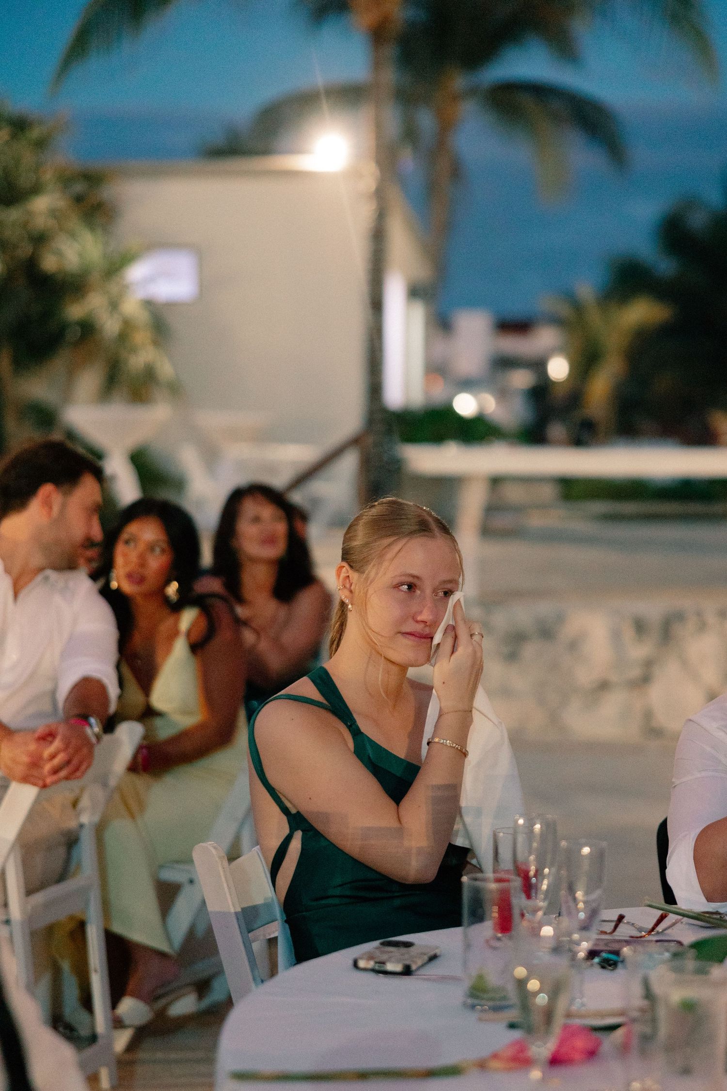 A woman in a green dress wipes a tear at an outdoor evening wedding reception near the beach.