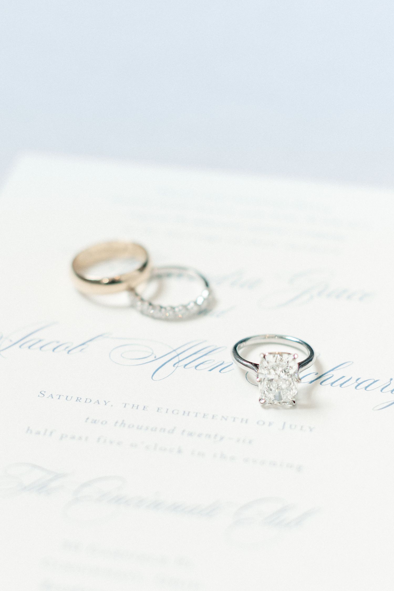 Elegant wedding rings and diamond engagement ring resting on a formal wedding invitation with blue script calligraphy.