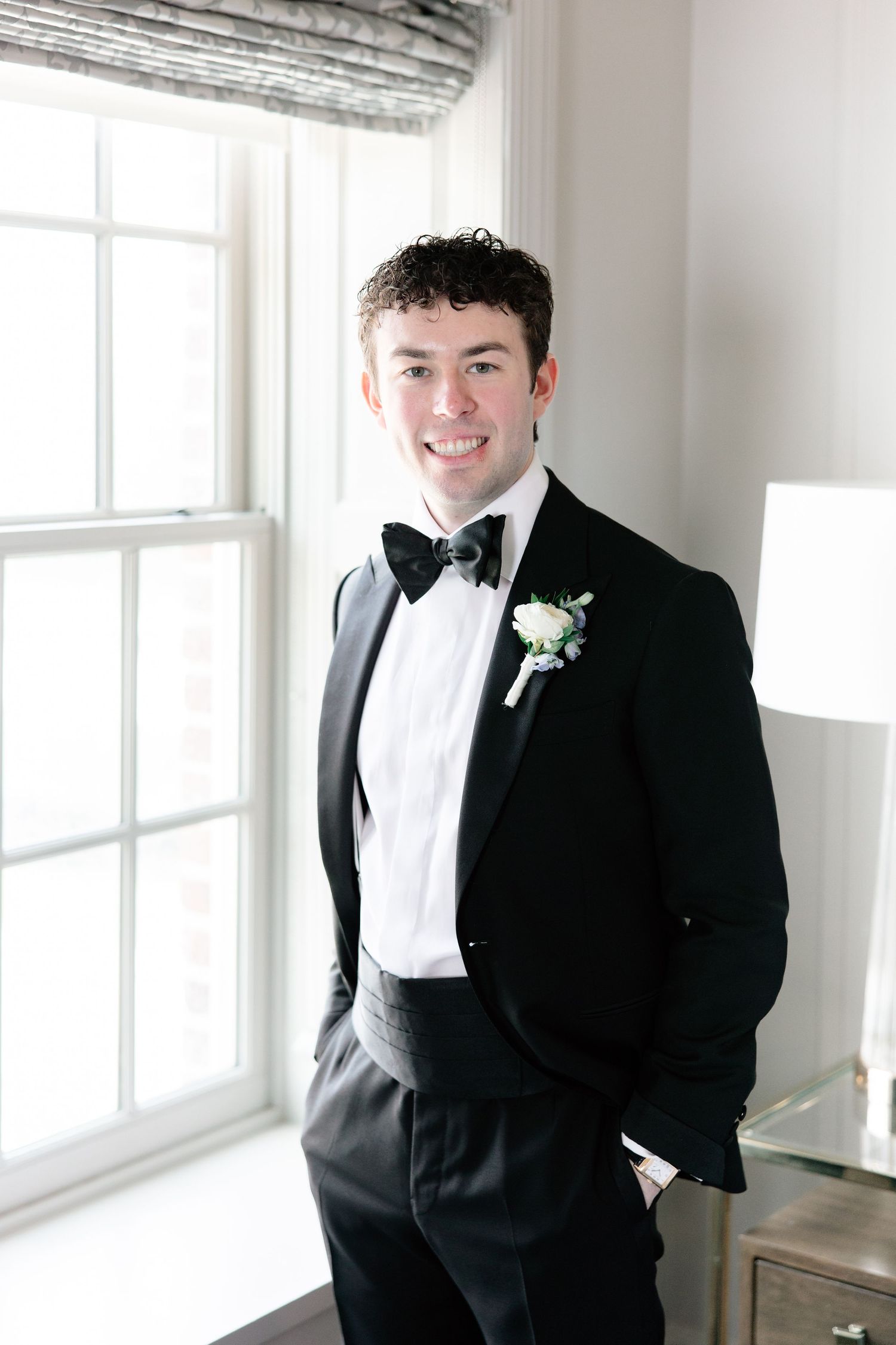 Smiling groom in black tuxedo with bow tie and white boutonniere stands by bright window on wedding day.