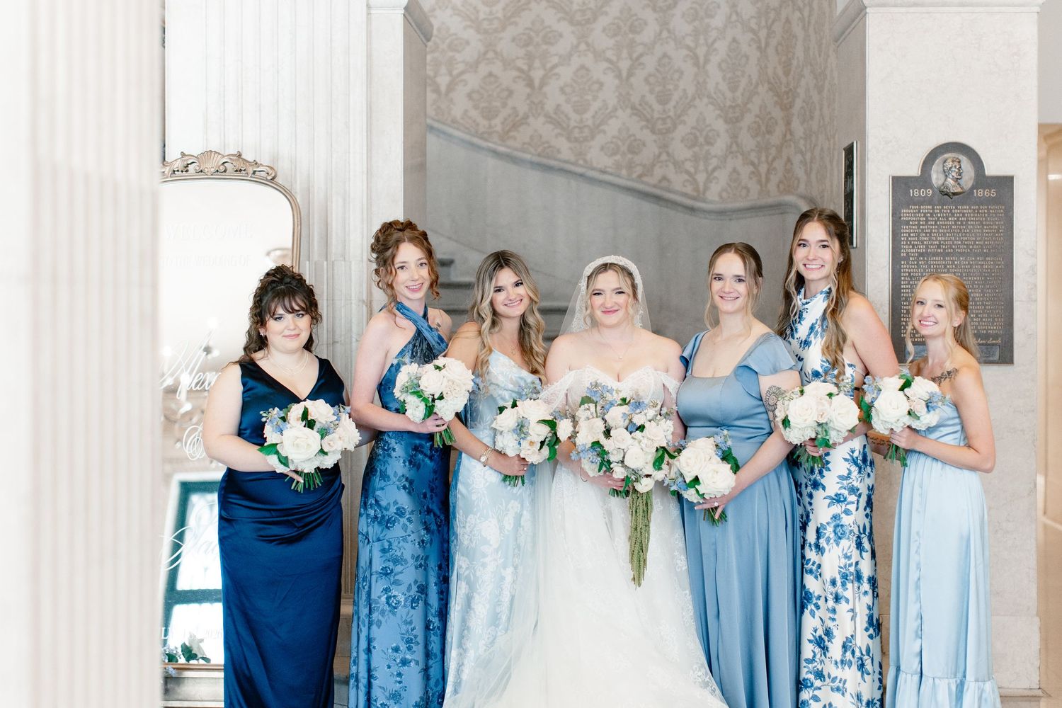 Bride in white gown poses with six bridesmaids in blue dresses holding white bouquets in elegant venue.