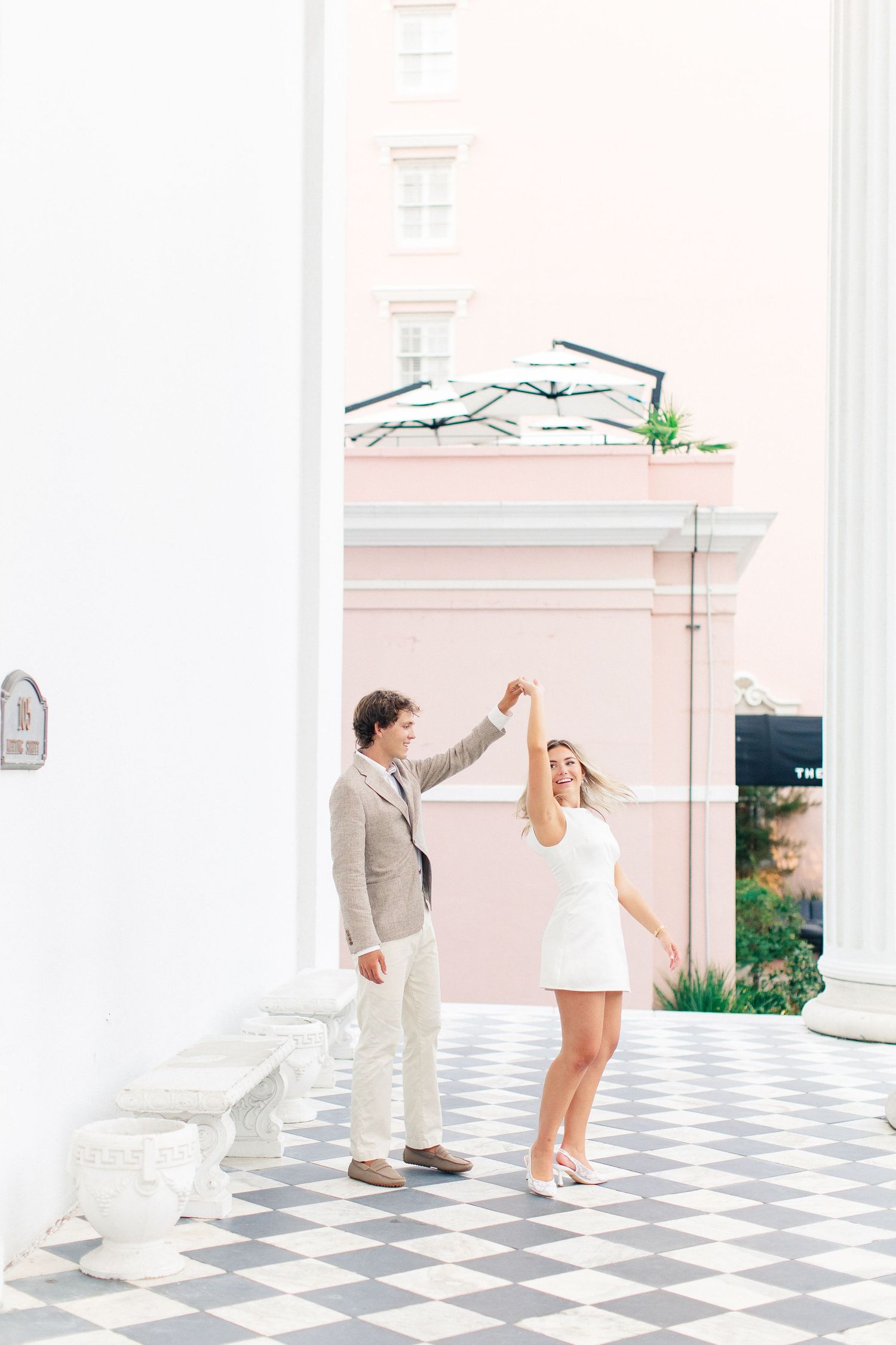 Couple dancing on checkered floor outdoors, man in tan suit spinning woman in white mini dress near pink building.