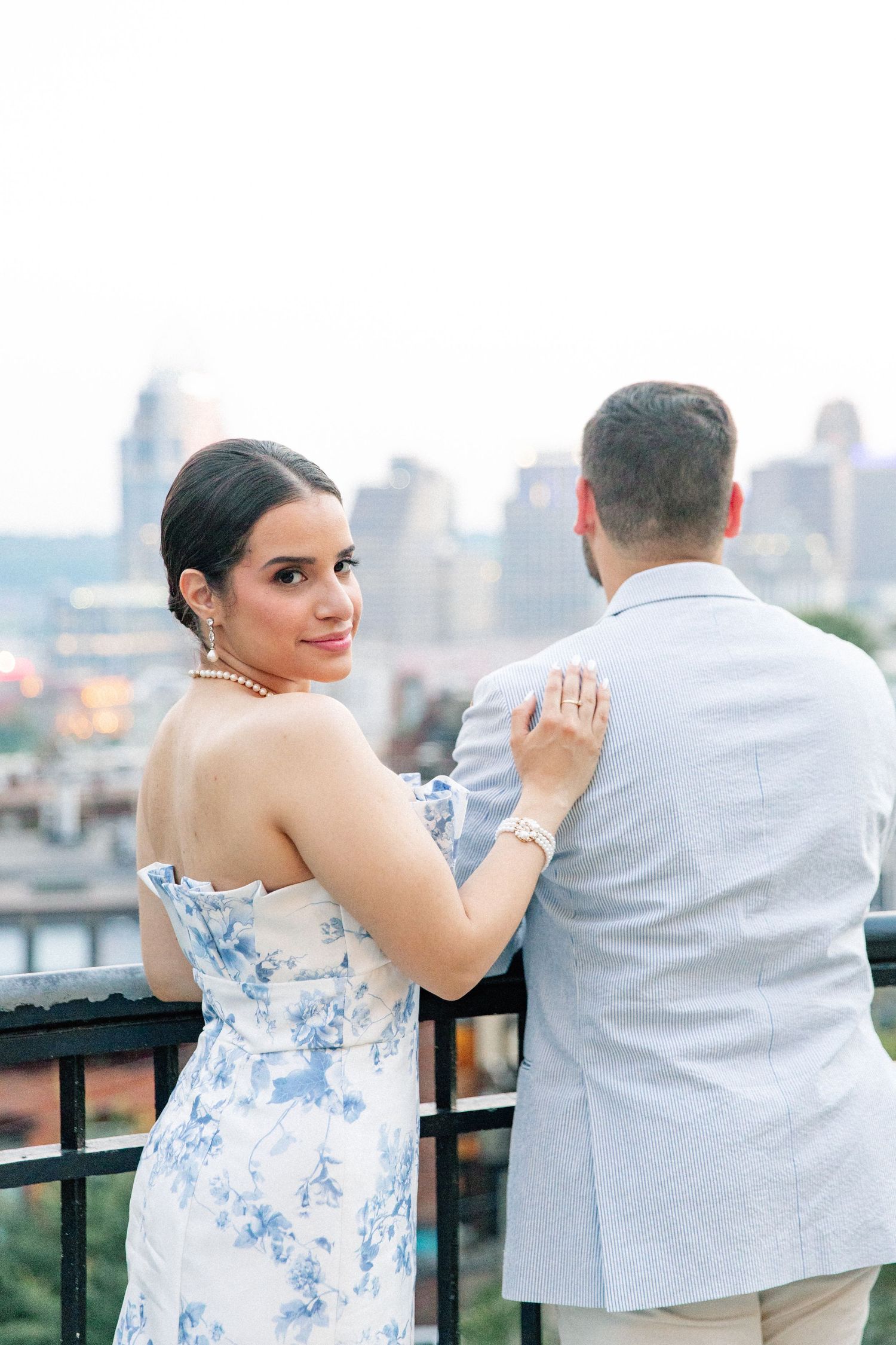 Bride in a blue floral gown looks back at the camera while standing with groom on a rooftop city overlook.