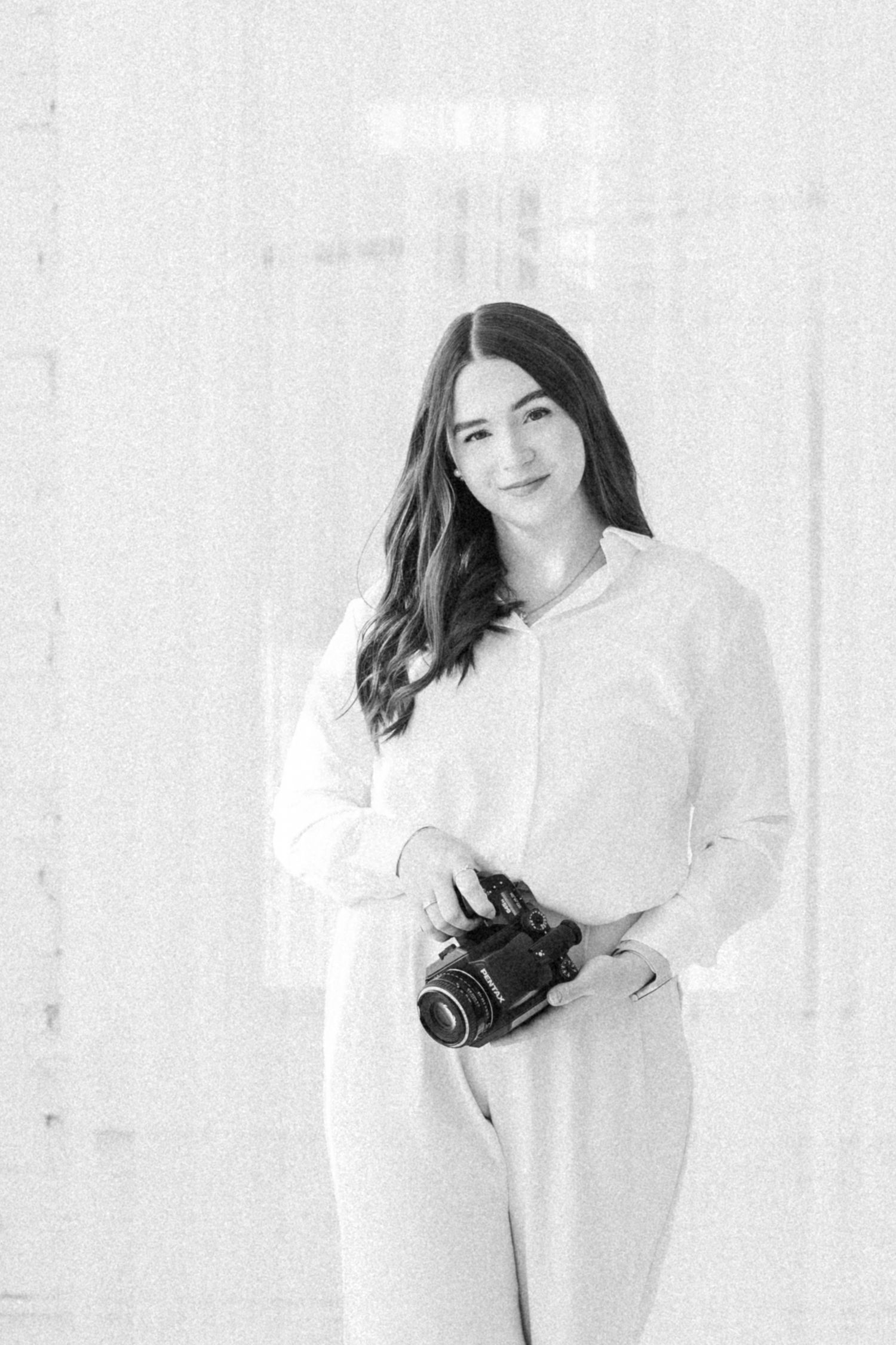 A smiling woman in a white blouse holds a DSLR camera in a softly lit, minimalist studio setting.
