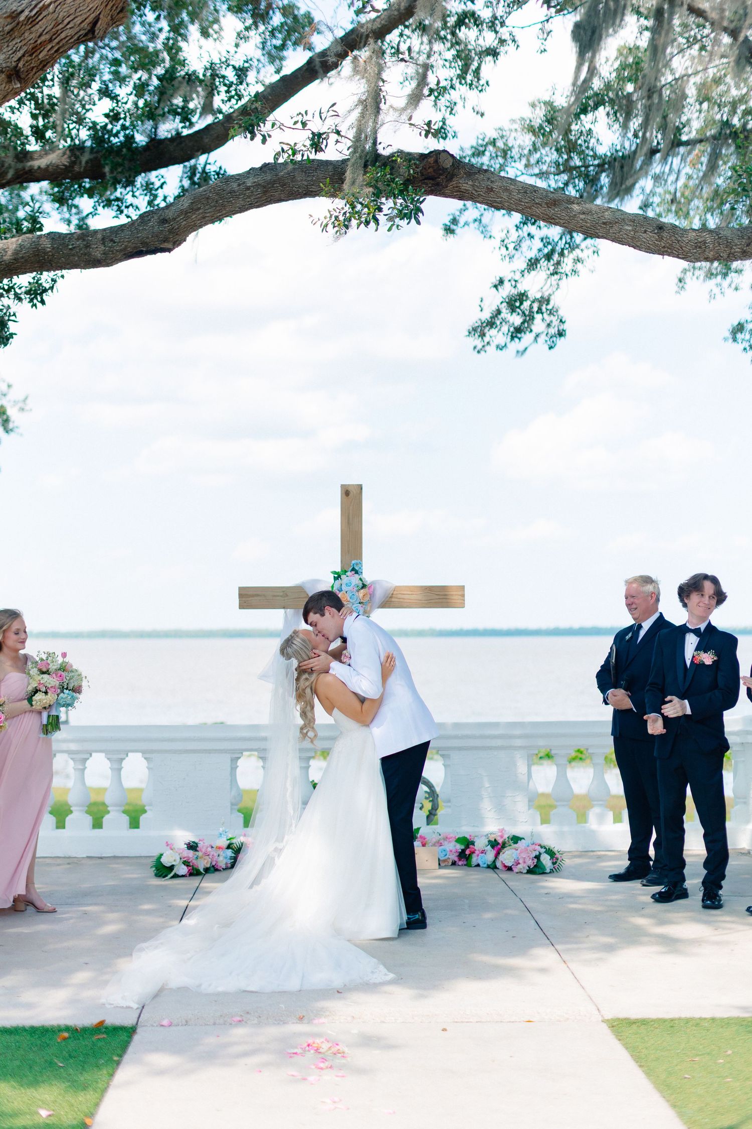 Bride and groom share first kiss at outdoor waterfront wedding ceremony beneath oak tree with wooden cross, wedding at Bella Cosa