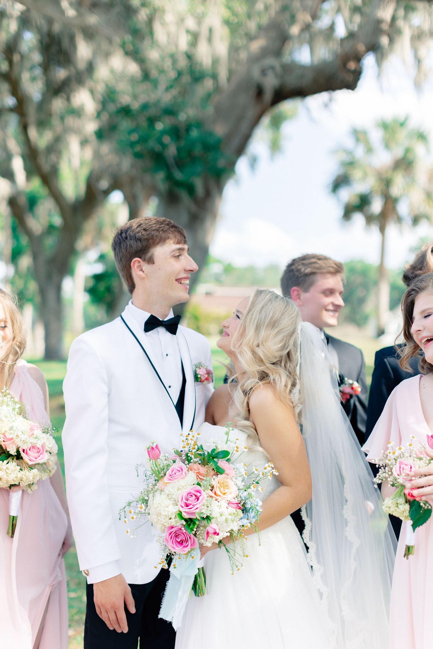Bride and groom smile together outdoors, surrounded by bridesmaids in pink holding bouquets of roses under Spanish moss trees, Bella Cosa