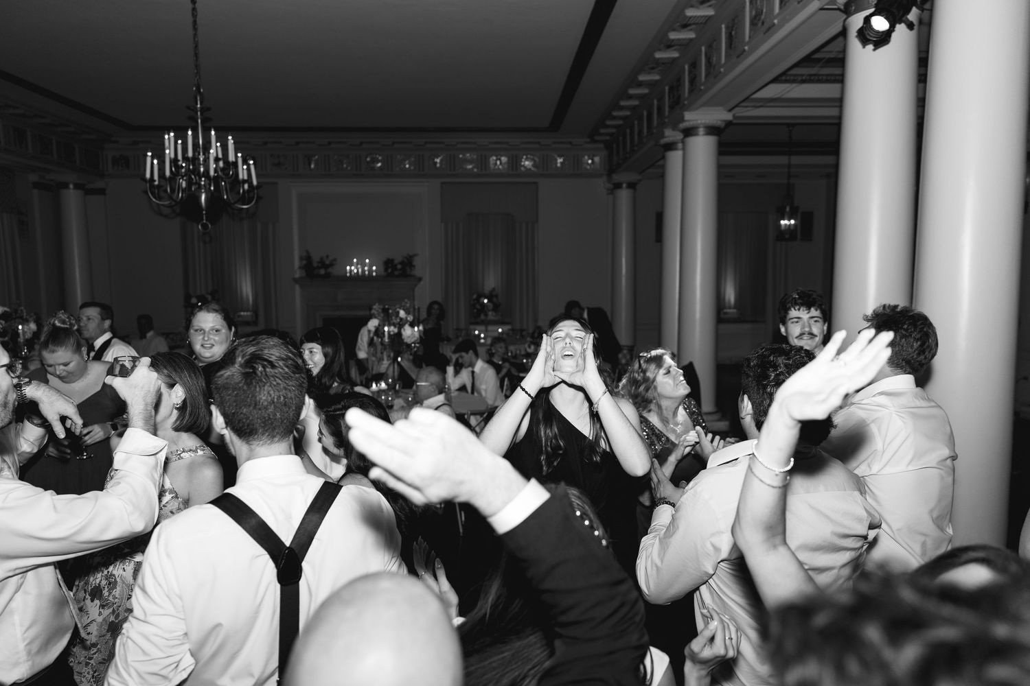 Black and white photo of wedding guests dancing and celebrating on a crowded dance floor in an elegant ballroom.
