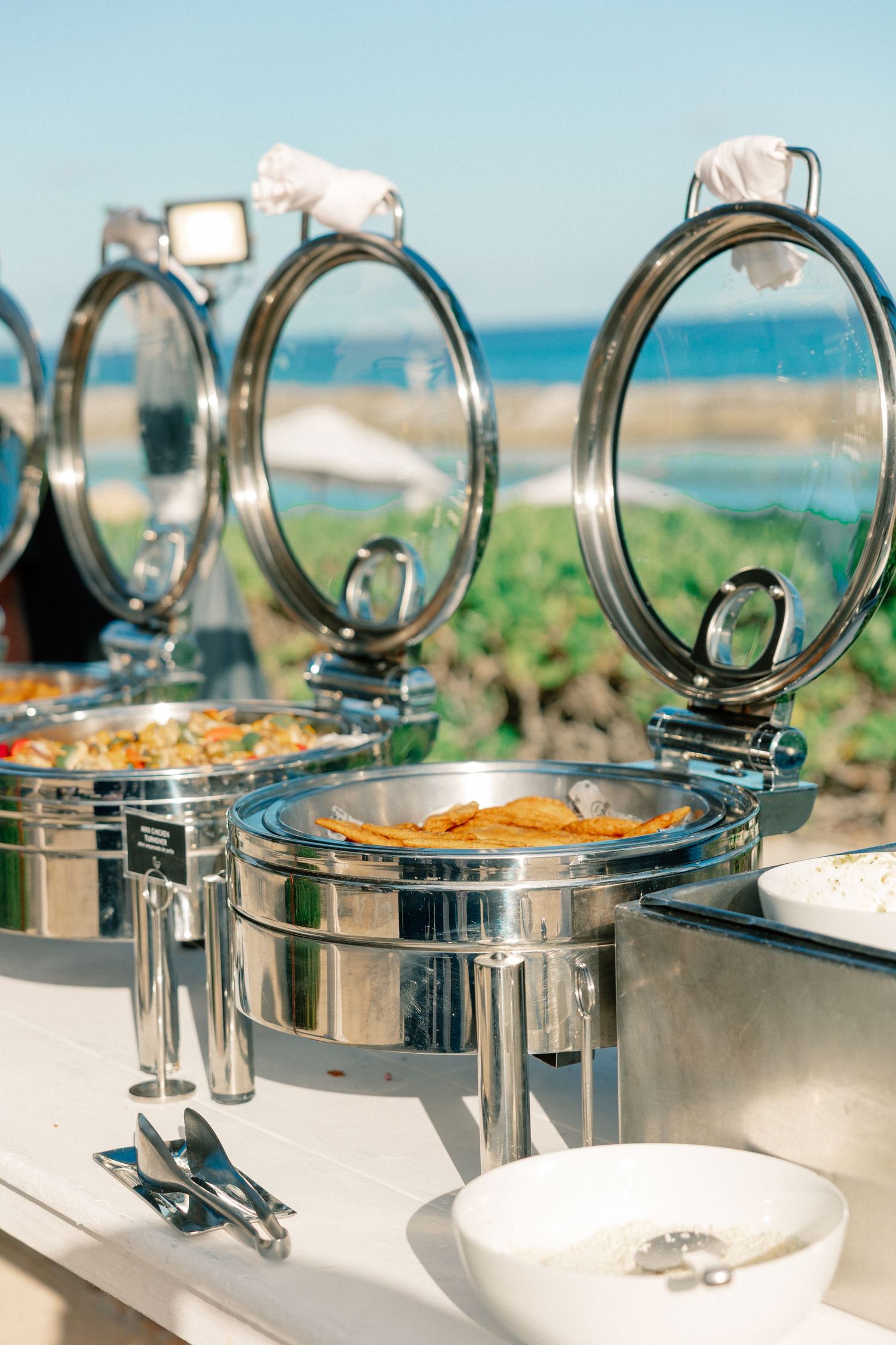 Stainless steel chafing dishes with open lids on a buffet table with an ocean view backdrop.
