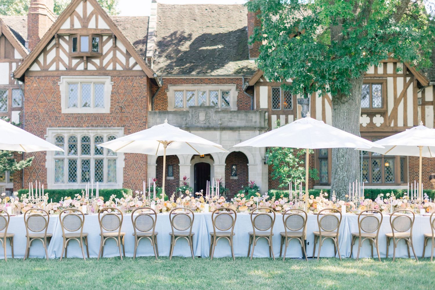 Elegant outdoor wedding reception with long tables, gold chairs, white umbrellas, and floral centerpieces at a Tudor-style manor.