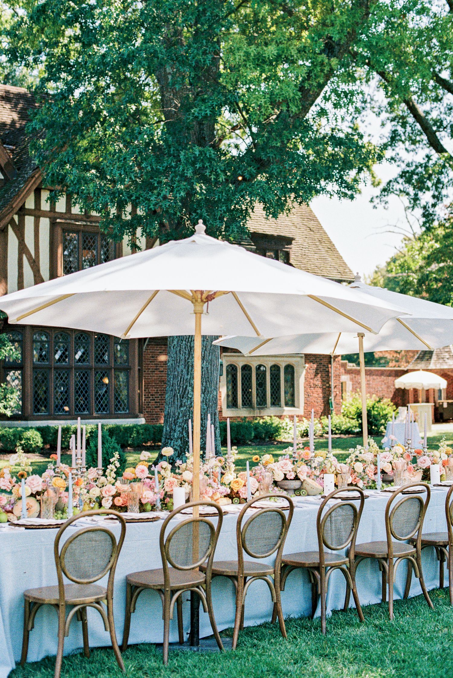 Elegant outdoor wedding reception with cane chairs, floral centerpieces, white umbrellas, and long tables on a manicured lawn.