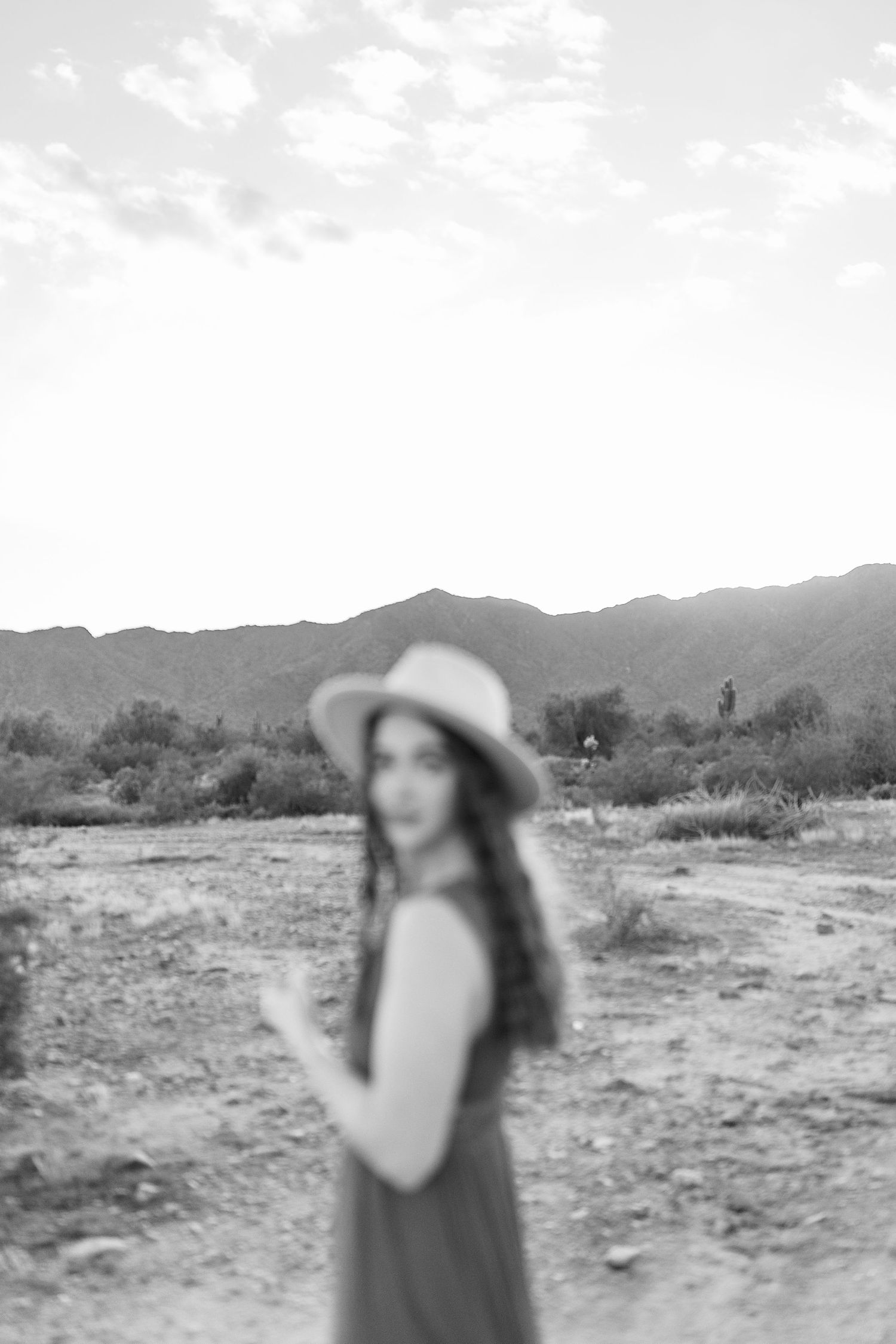 Black and white portrait of a woman in a wide brim hat standing in a desert landscape with mountains.