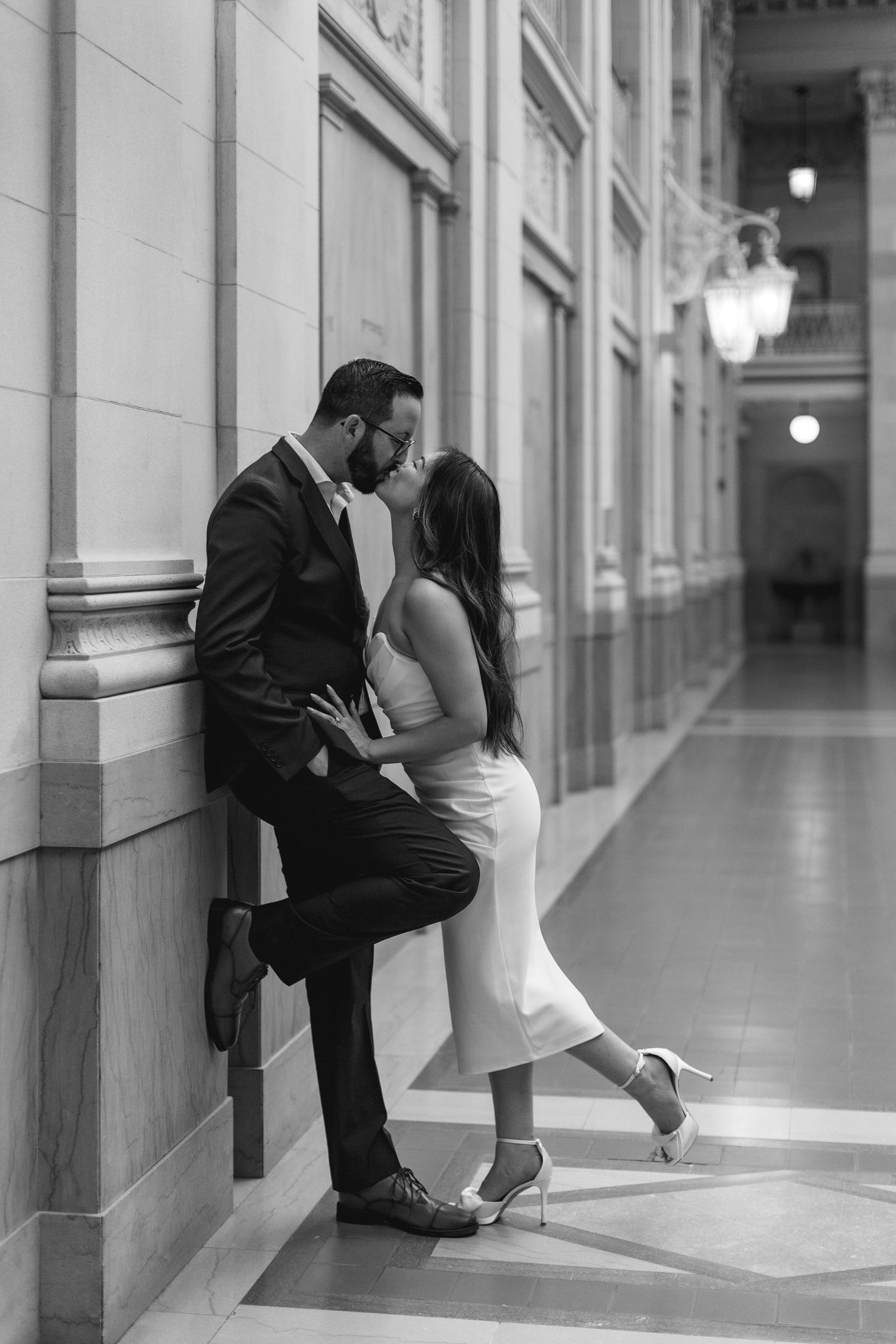 Black and white photo of a couple sharing a romantic kiss in an elegant hallway, the bride in a white dress and groom in a suit.