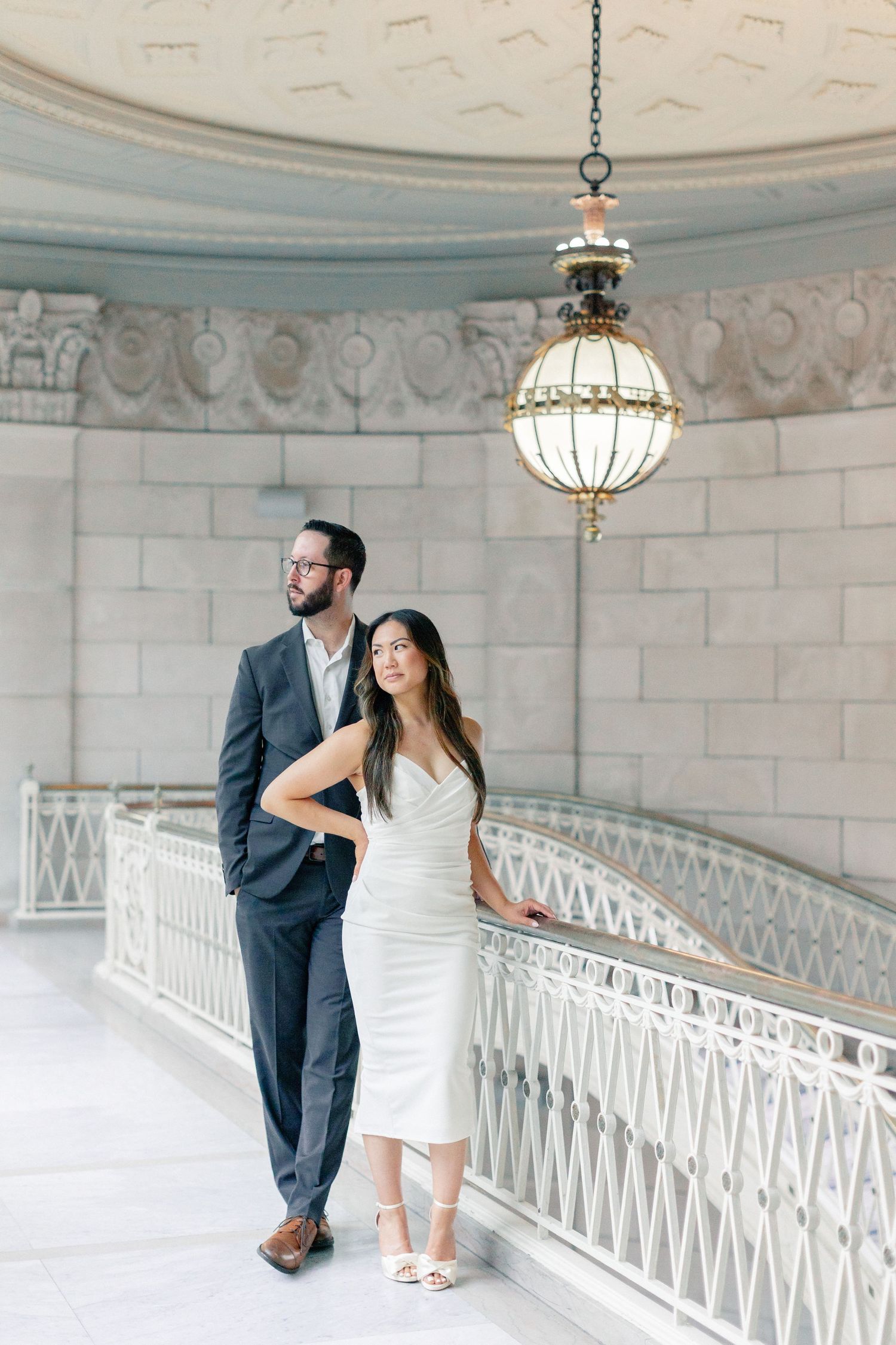 Elegant couple posing on a grand staircase beneath a gold chandelier in a classical marble interior.