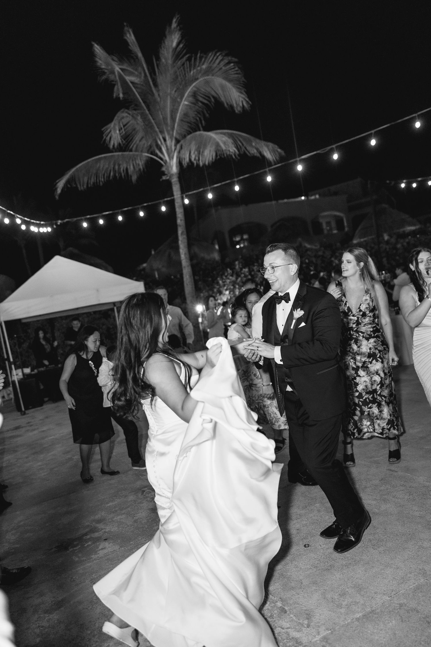 Black and white photo of bride and groom dancing outdoors at night under string lights with palm tree backdrop.