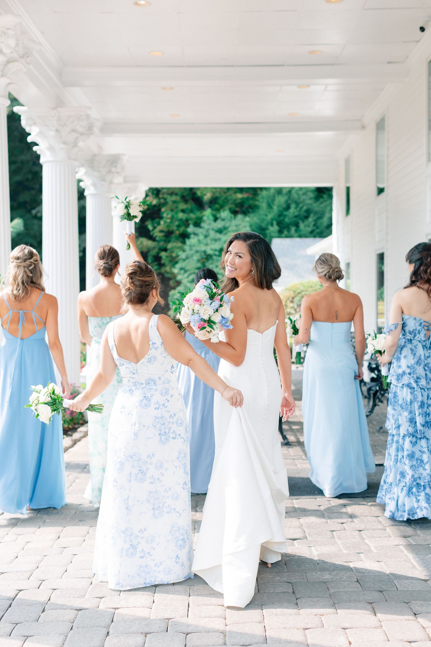 Bride in white gown smiles holding bouquet with bridesmaids in blue dresses walking along elegant columned venue.