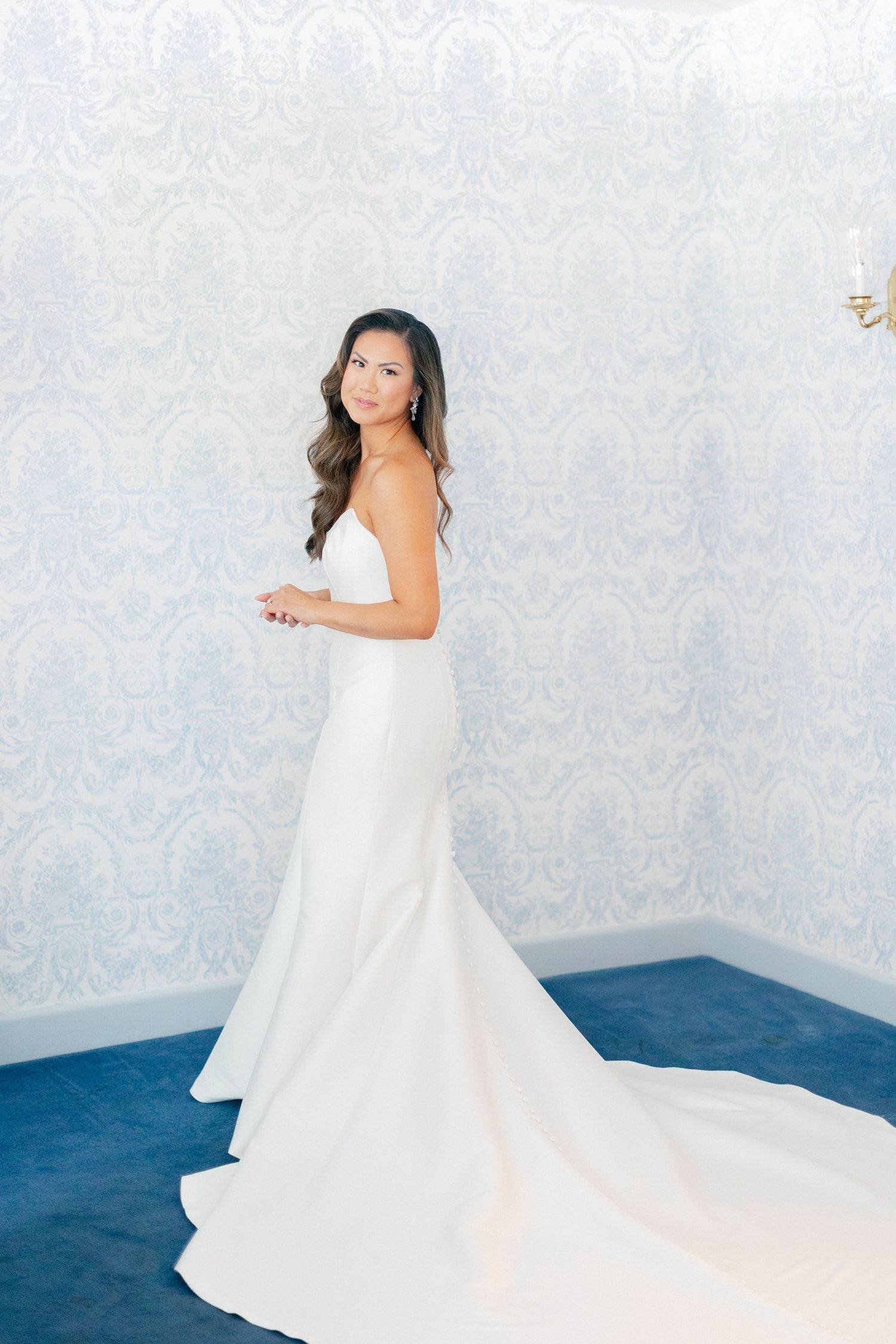 Elegant bride in a sleek white satin mermaid wedding gown with a train, posing in a blue wallpapered room.