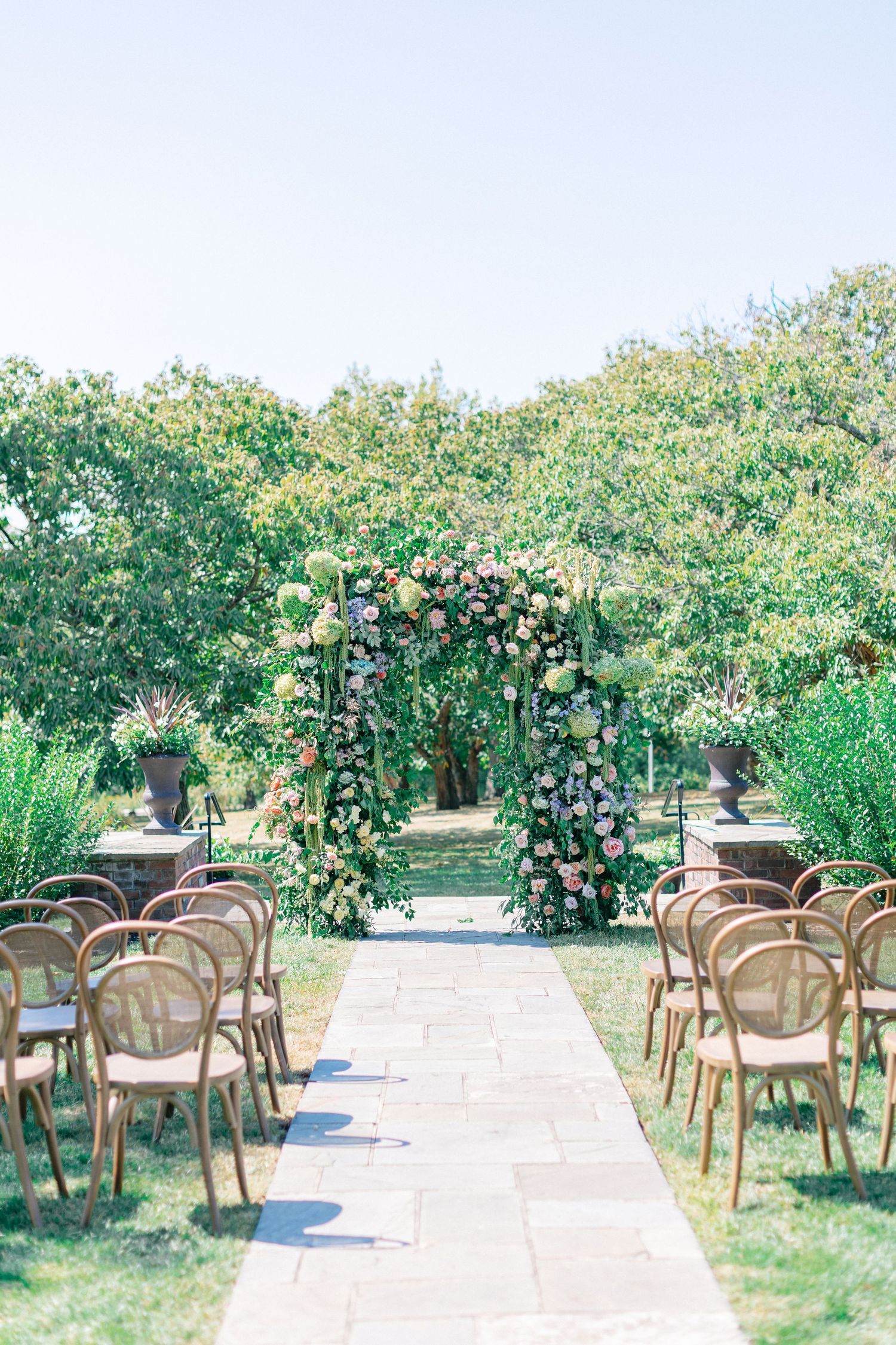 Outdoor wedding ceremony setup with floral arch, wooden chairs lining stone aisle, surrounded by lush greenery under clear blue sky.
