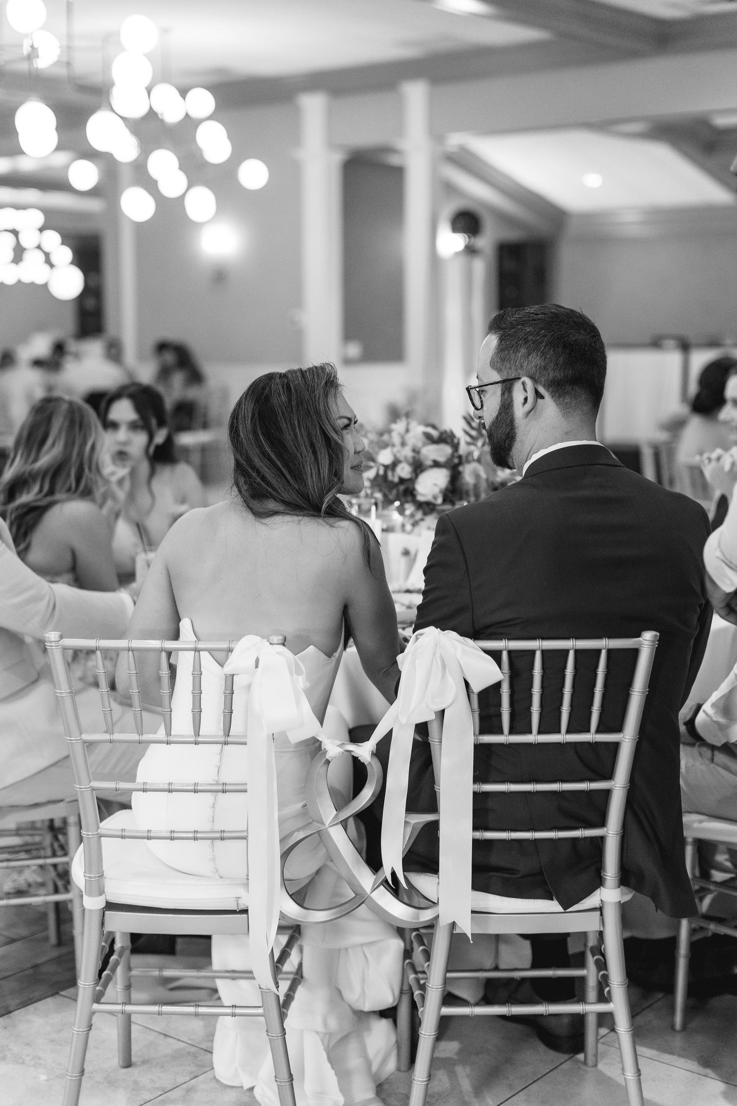 Black and white photo of bride and groom seated at wedding reception table, chairs adorned with elegant ribbon bows.