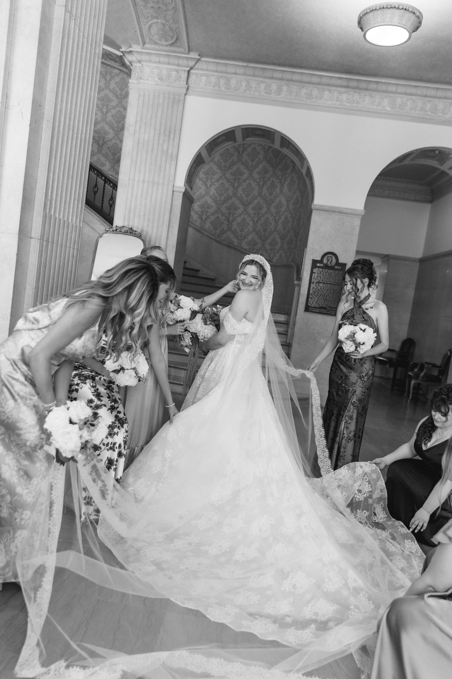Bridesmaids helping bride in elegant lace wedding gown and veil in ornate venue, black and white photo.