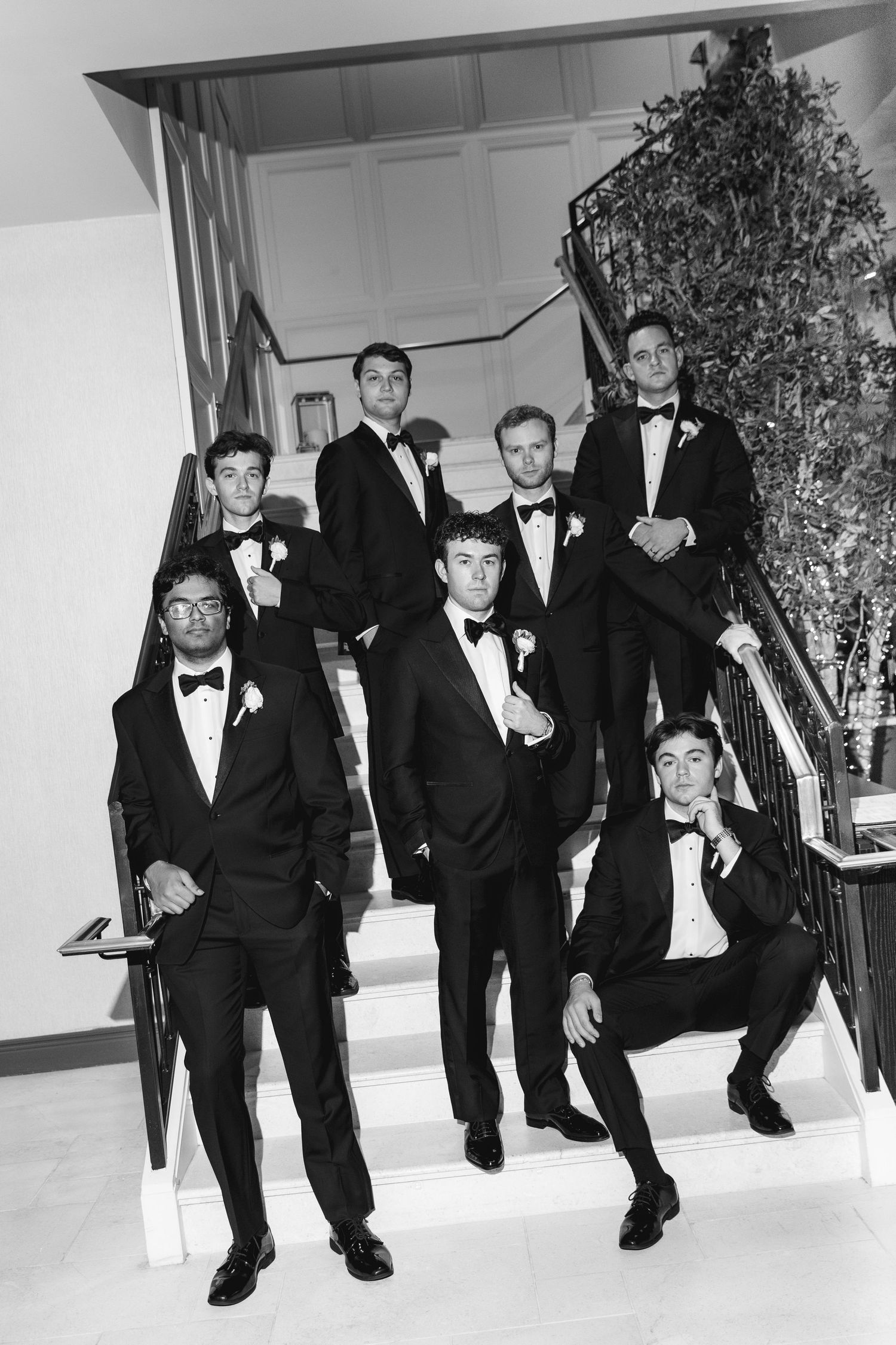Black and white photo of seven groomsmen in tuxedos posing on an elegant staircase at a wedding.
