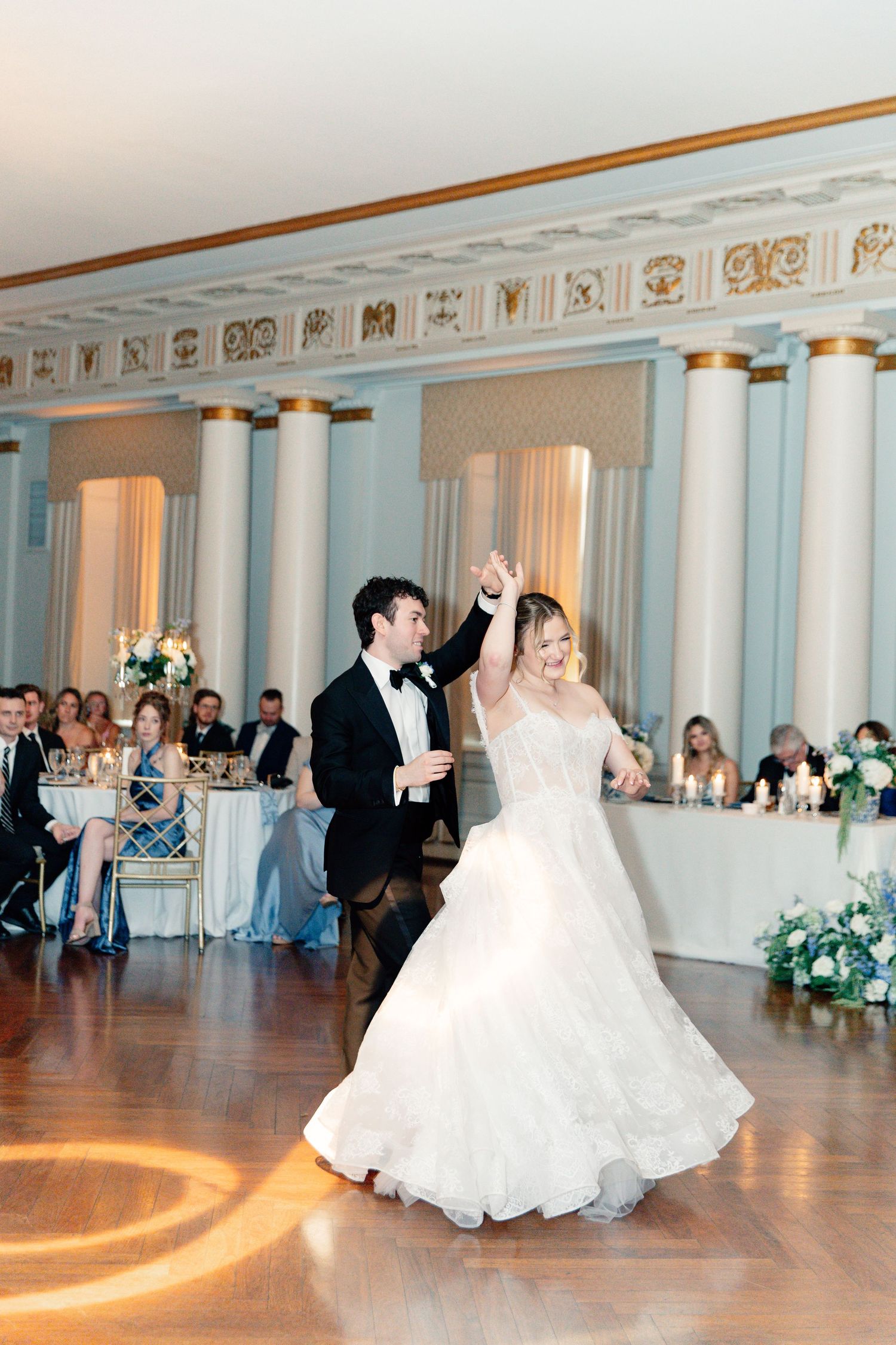 Bride and groom share their first dance in an elegant ballroom with white columns and gold accents at their wedding reception.