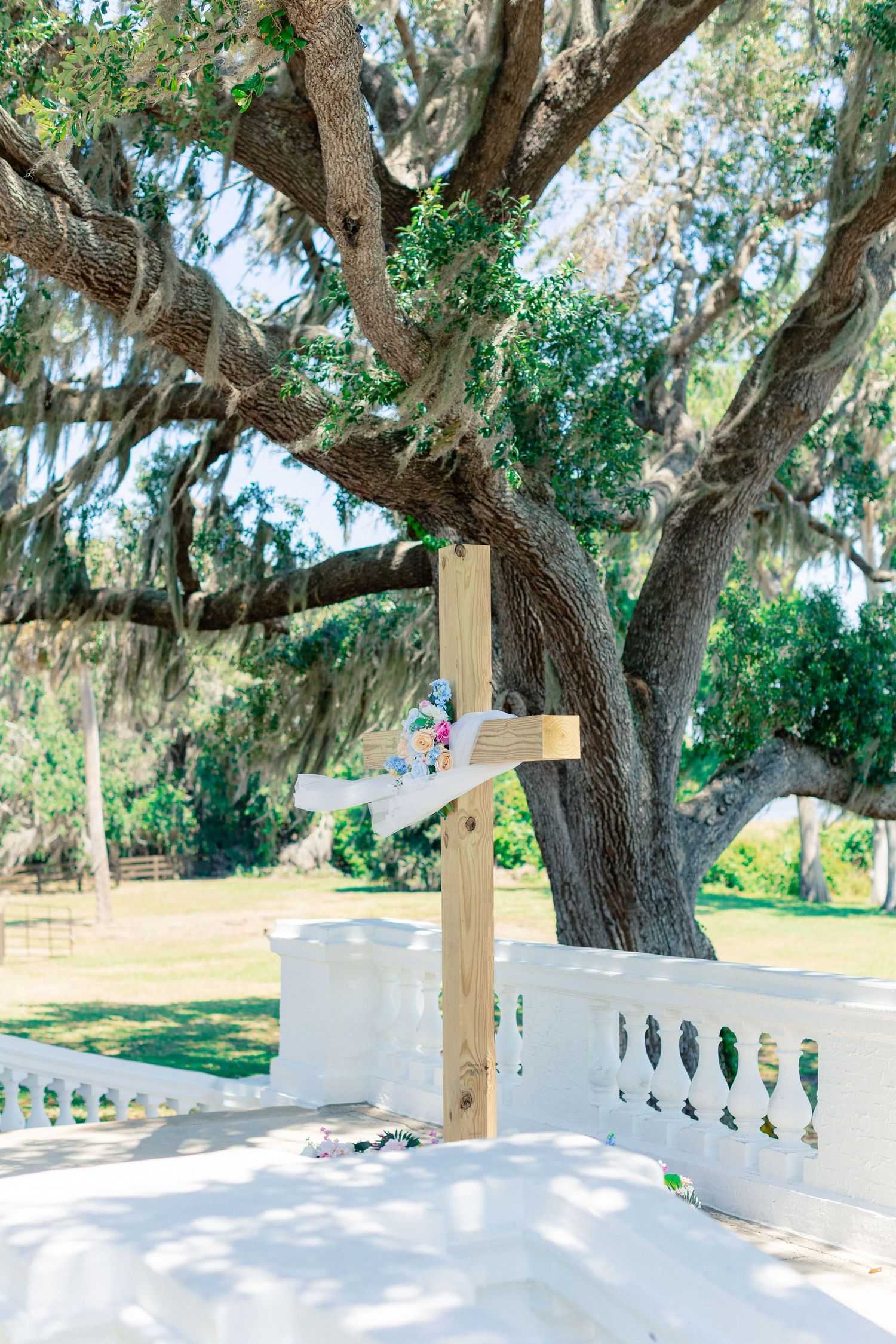 Wooden cross decorated with flowers and ribbon stands before a grand oak tree at an outdoor wedding ceremony, wedding at Bella Cosa