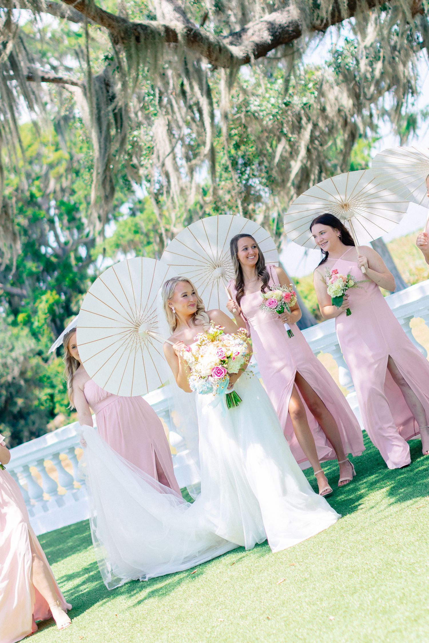 Bride in white gown walks with bridesmaids in pink dresses holding parasols and bouquets outdoors, wedding at Bella Cosa