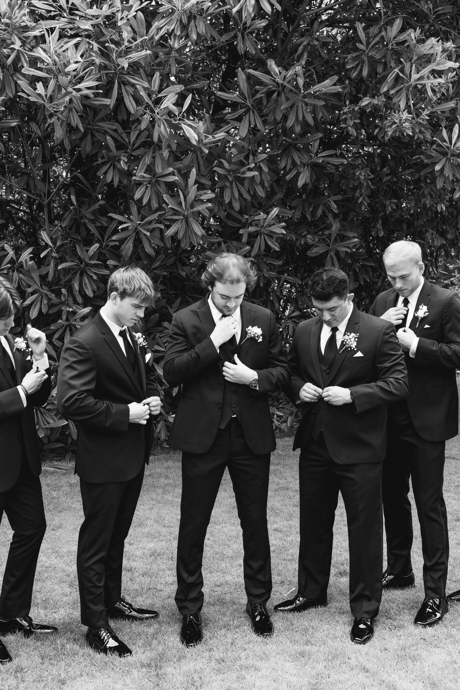 Groomsmen in black suits adjusting boutonnieres outdoors before a wedding ceremony.