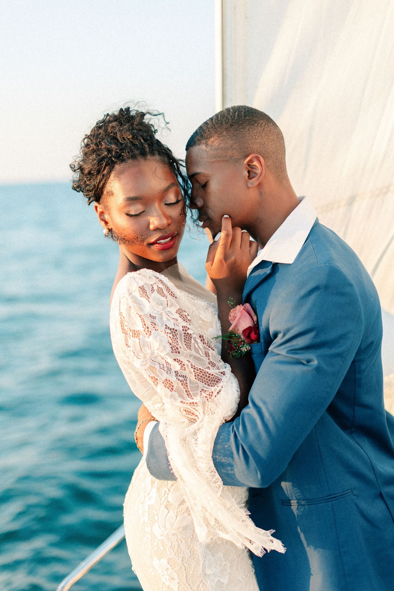 Elegant Black couple in wedding attire embrace romantically on a sailboat with ocean backdrop.