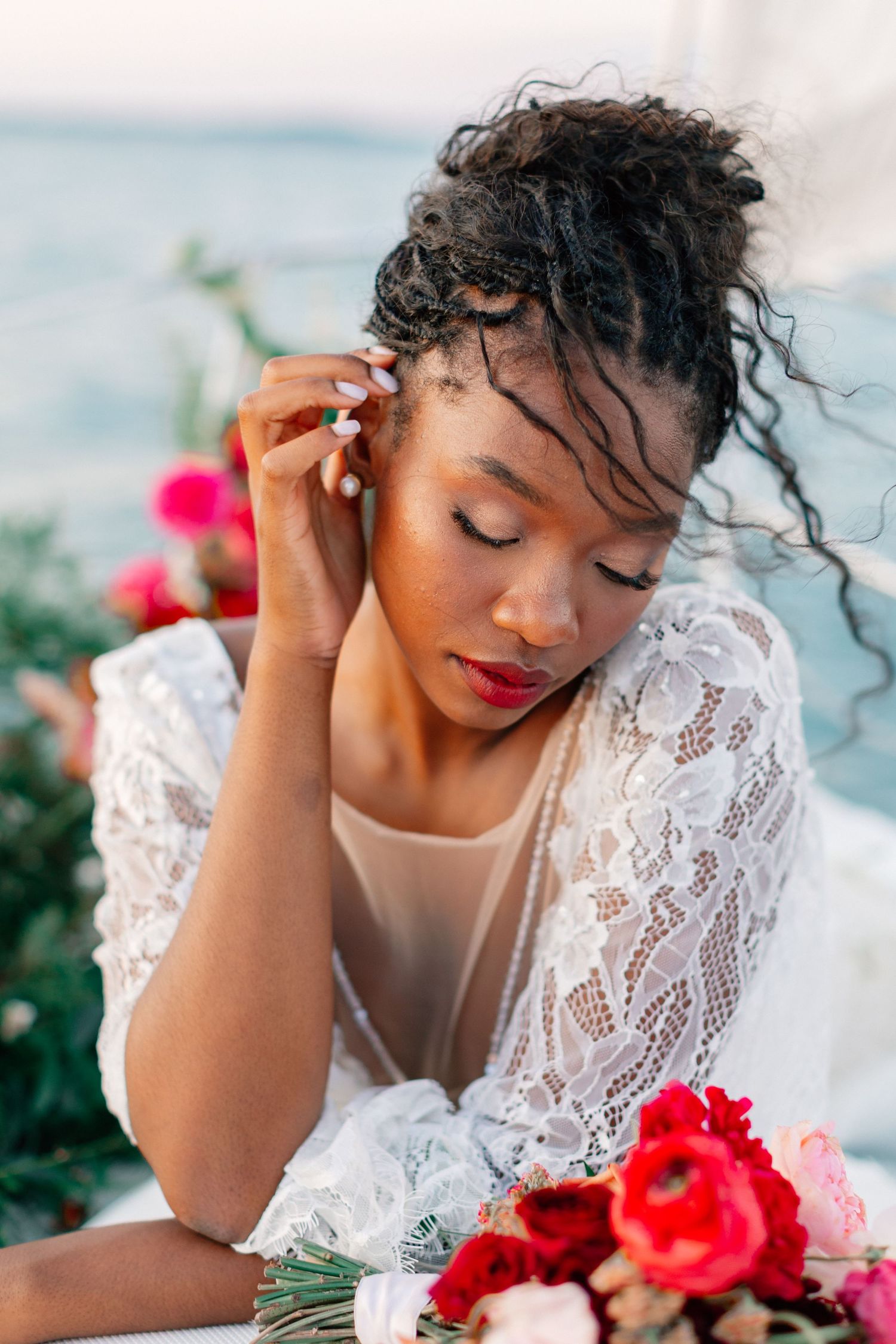 Black bride in white lace dress adjusting earring with red floral bouquet and ocean backdrop.